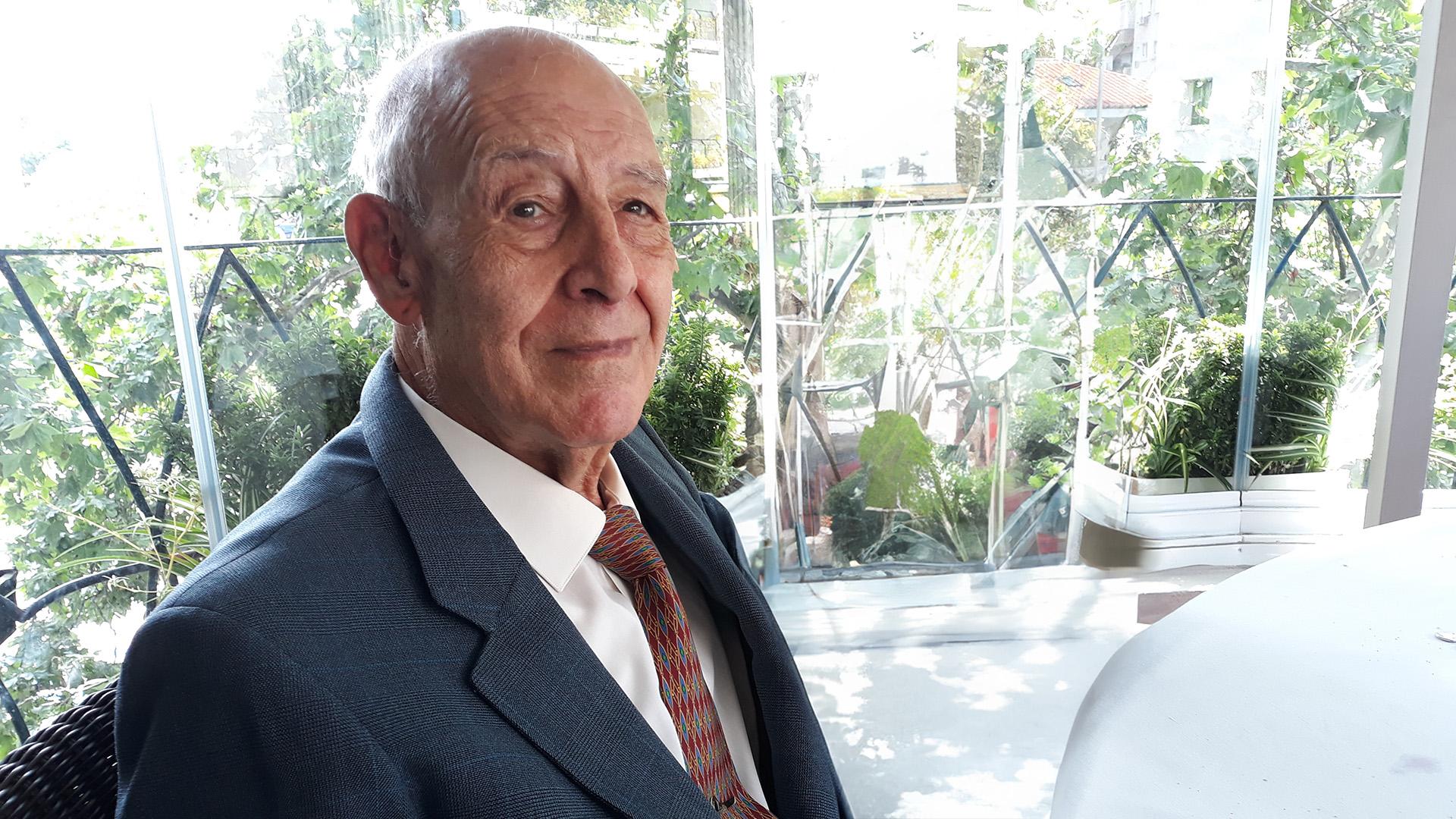 An elderly man in a suit and striped tie smiling at the camera in a bright, plant-filled conservatory.