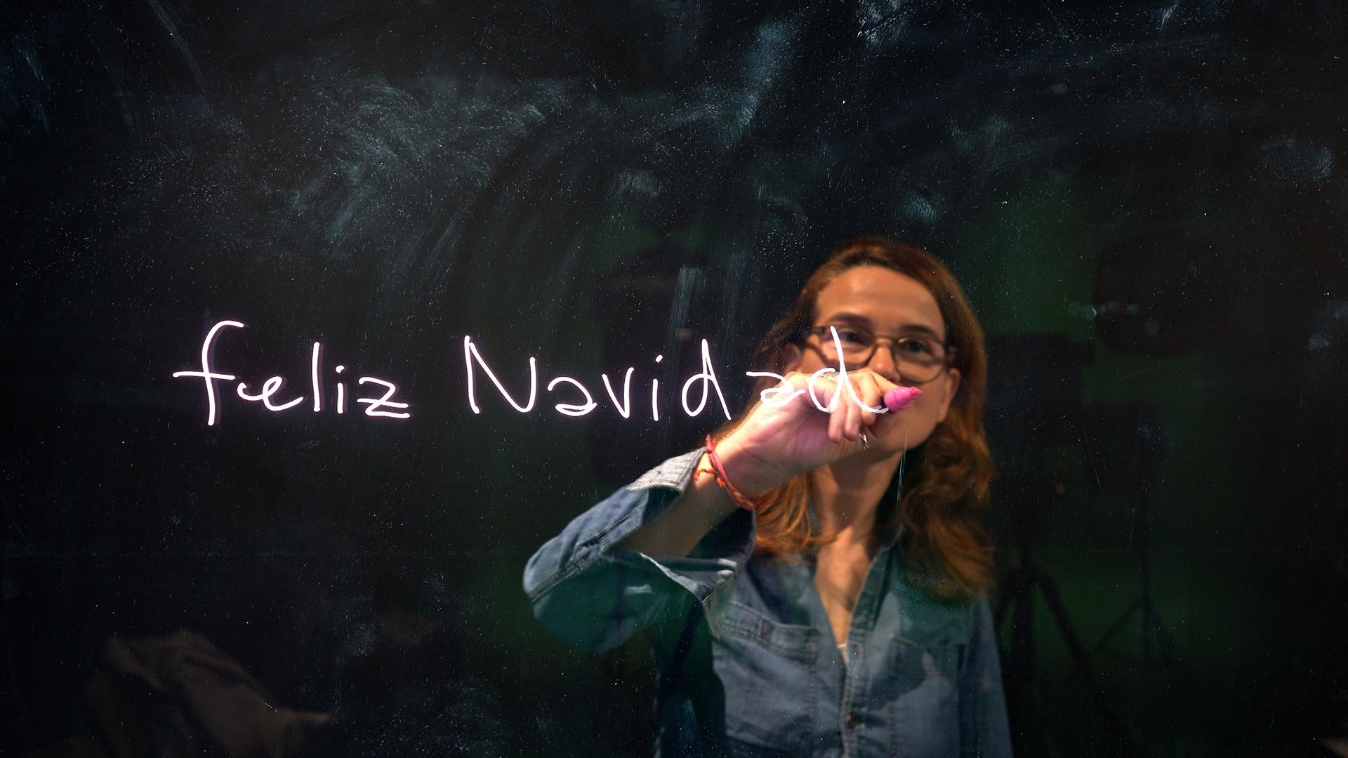 Una mujer escribe 'feliz Navidad' en un cristal con una luz brillante.