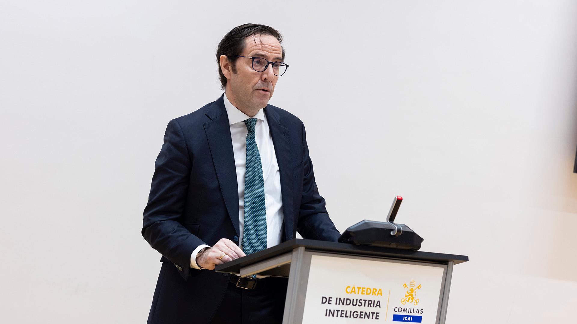 Un hombre vestido de traje se encuentra de pie frente a un micrófono, mientras habla durante un evento.