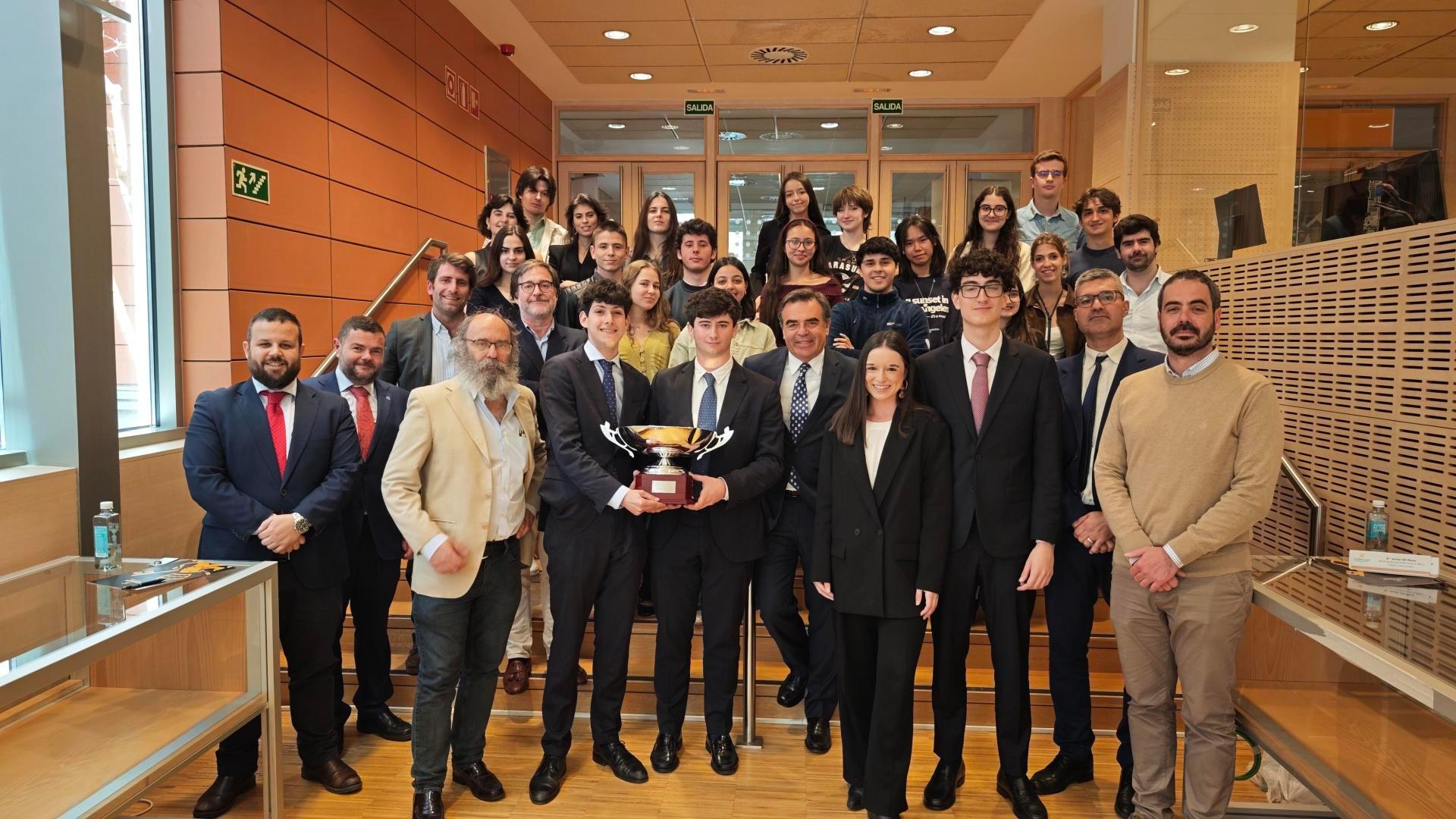 Un grupo de estudiantes y profesores posan juntos en un evento, sosteniendo un trofeo en el centro.