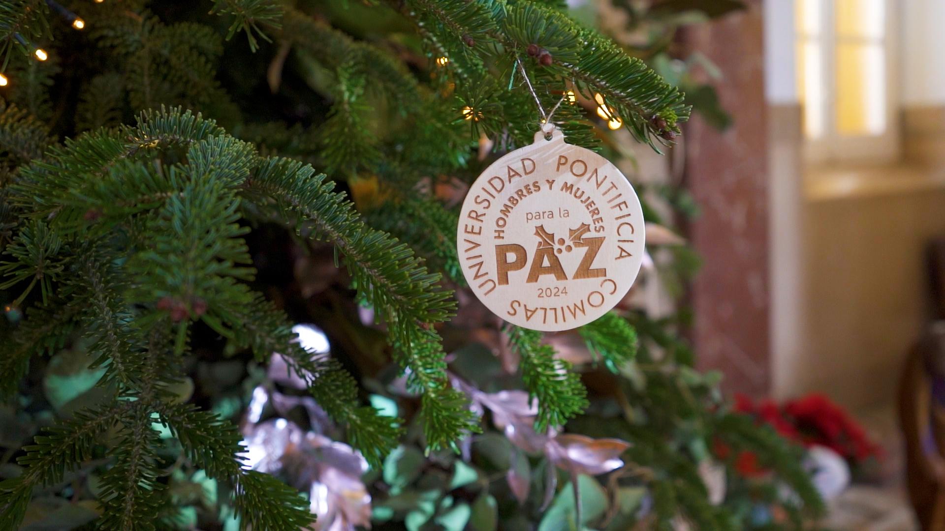 Un adorno navideño con el logo y texto de la Universidad Pontificia colgado en un árbol de Navidad iluminado.