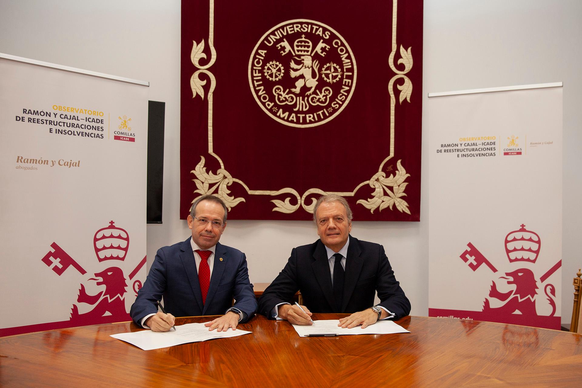 Two men in suits sitting at a table signing documents, with a red and gold university crest banner in the background.