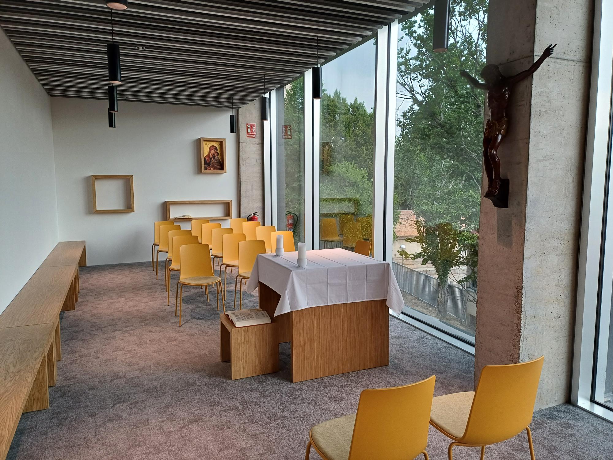 La imagen muestra un interior de una sala con bancos, mesas y un altar junto a grandes ventanales que permiten la entrada de luz natural.