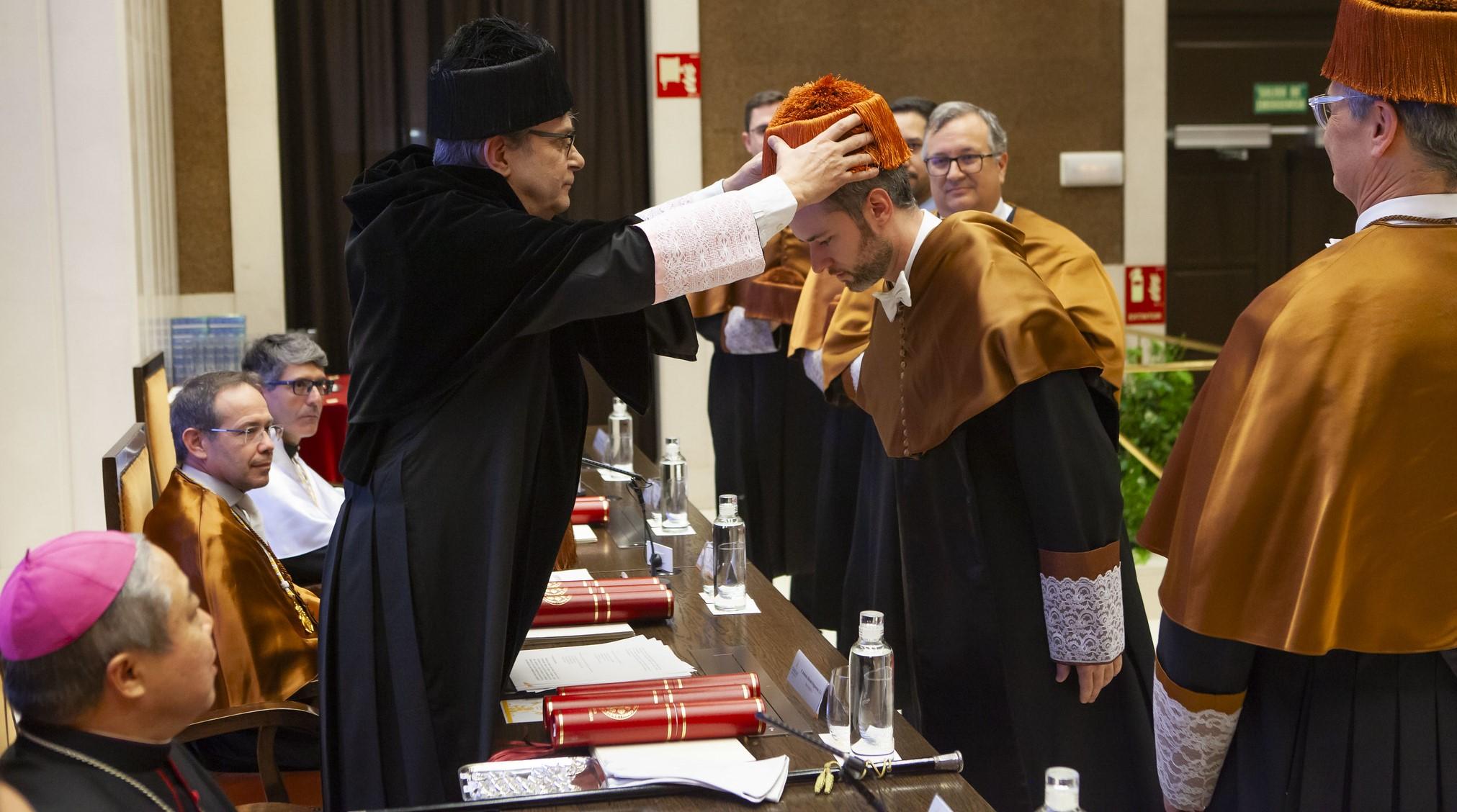 Un grupo de personas vestidas con togas y birretes participa en una ceremonia académica en un salón adornado con libros.