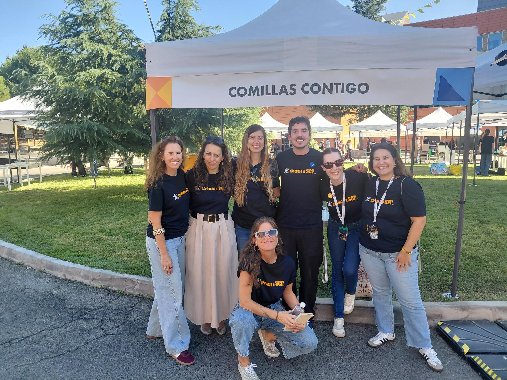 Un grupo de siete personas sonrientes posando frente a una carpa con el letrero 'COMILLAS CONTIGO'.