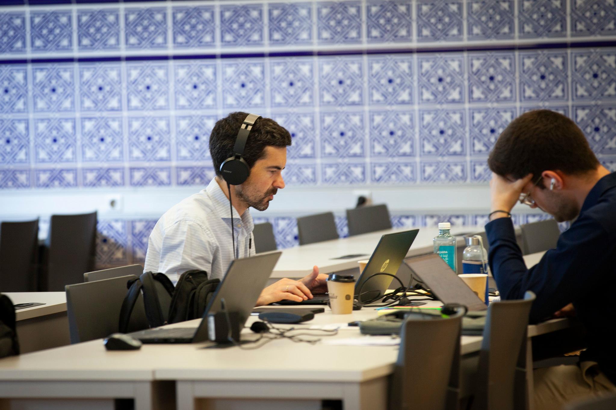 Two men working on laptops in a modern office space with blue tiled walls.