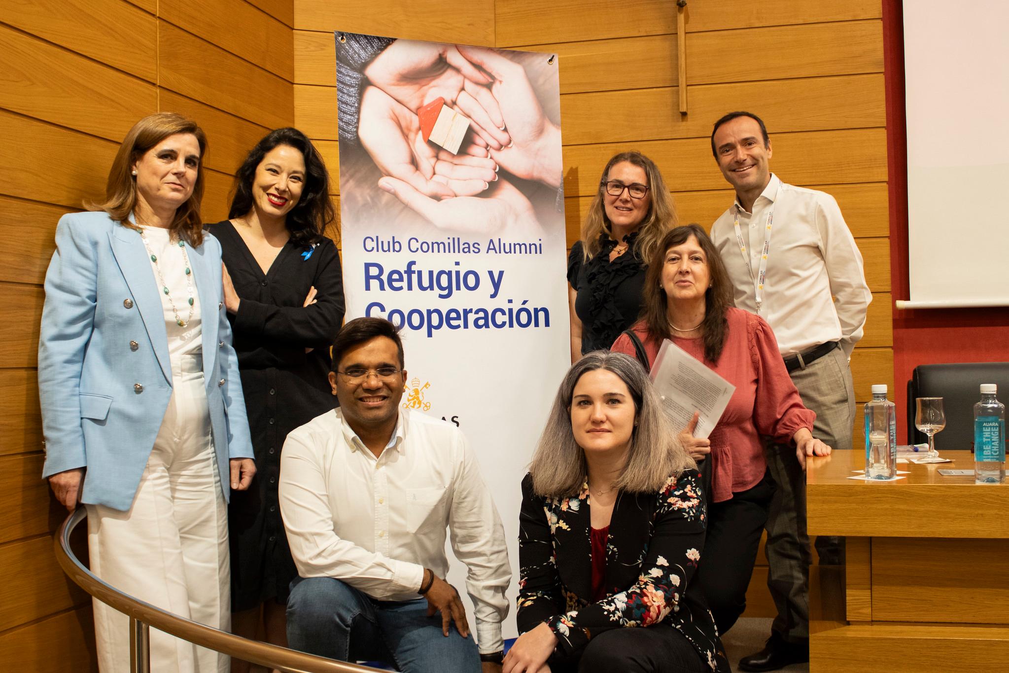 A group of seven people posing in front of a banner reading 'Club Comillas Alumni Refugio y Cooperación' inside a building.