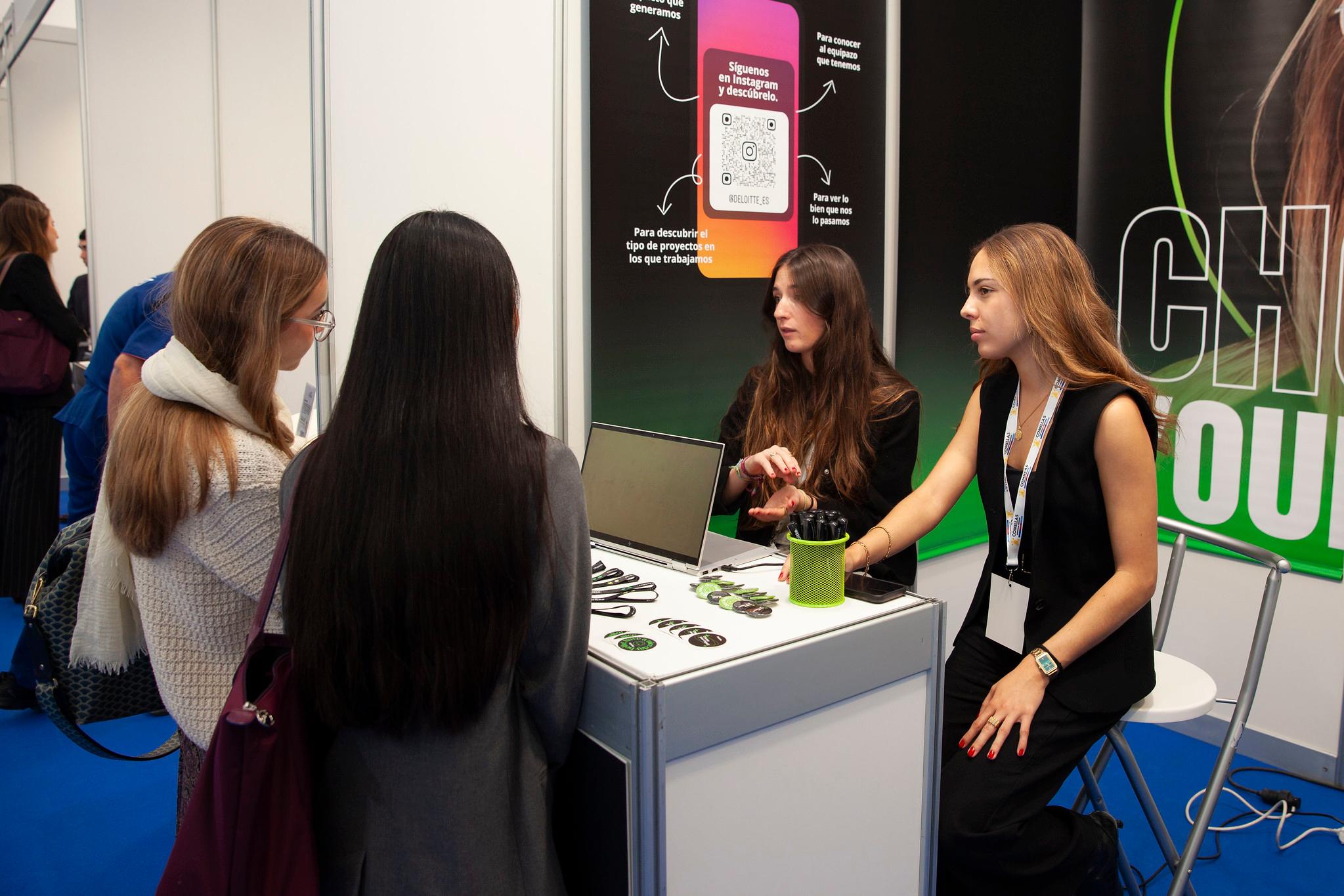 Four women engaged in a conversation at a promotional booth with laptops and brochures.