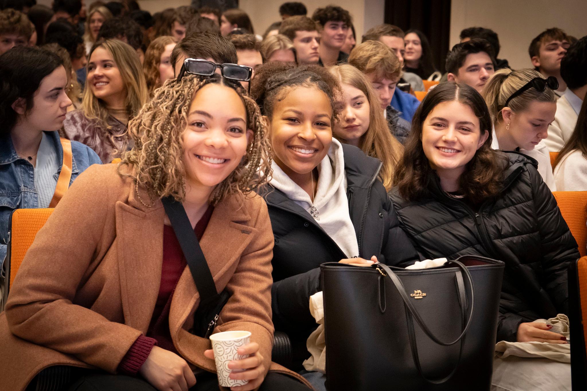 Tres jóvenes sonrientes sentadas entre la multitud en lo que parece ser un auditorio.