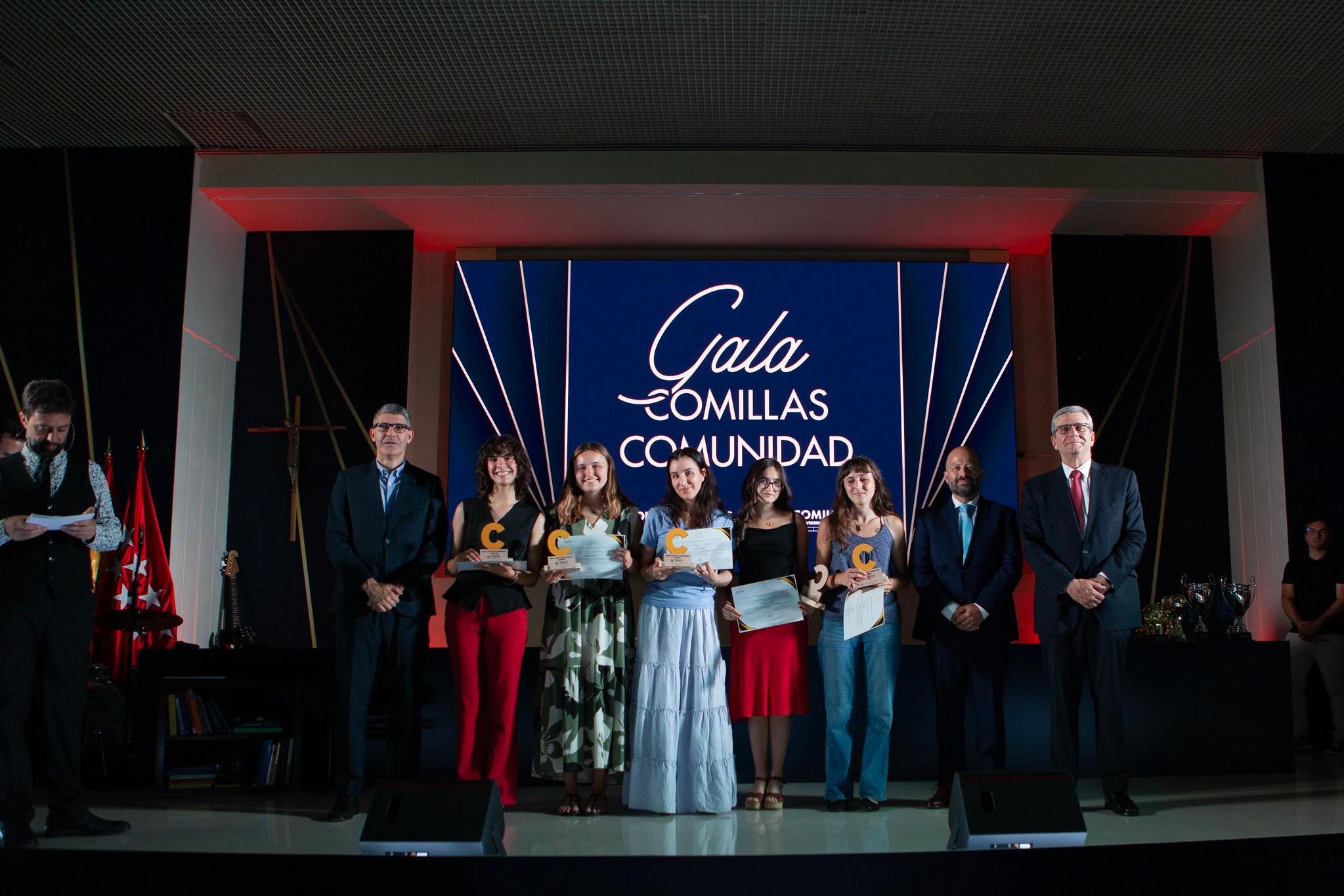 Un grupo de personas en un escenario durante la Gala Comillas Comunidad, sosteniendo reconocimientos frente a un fondo iluminado.