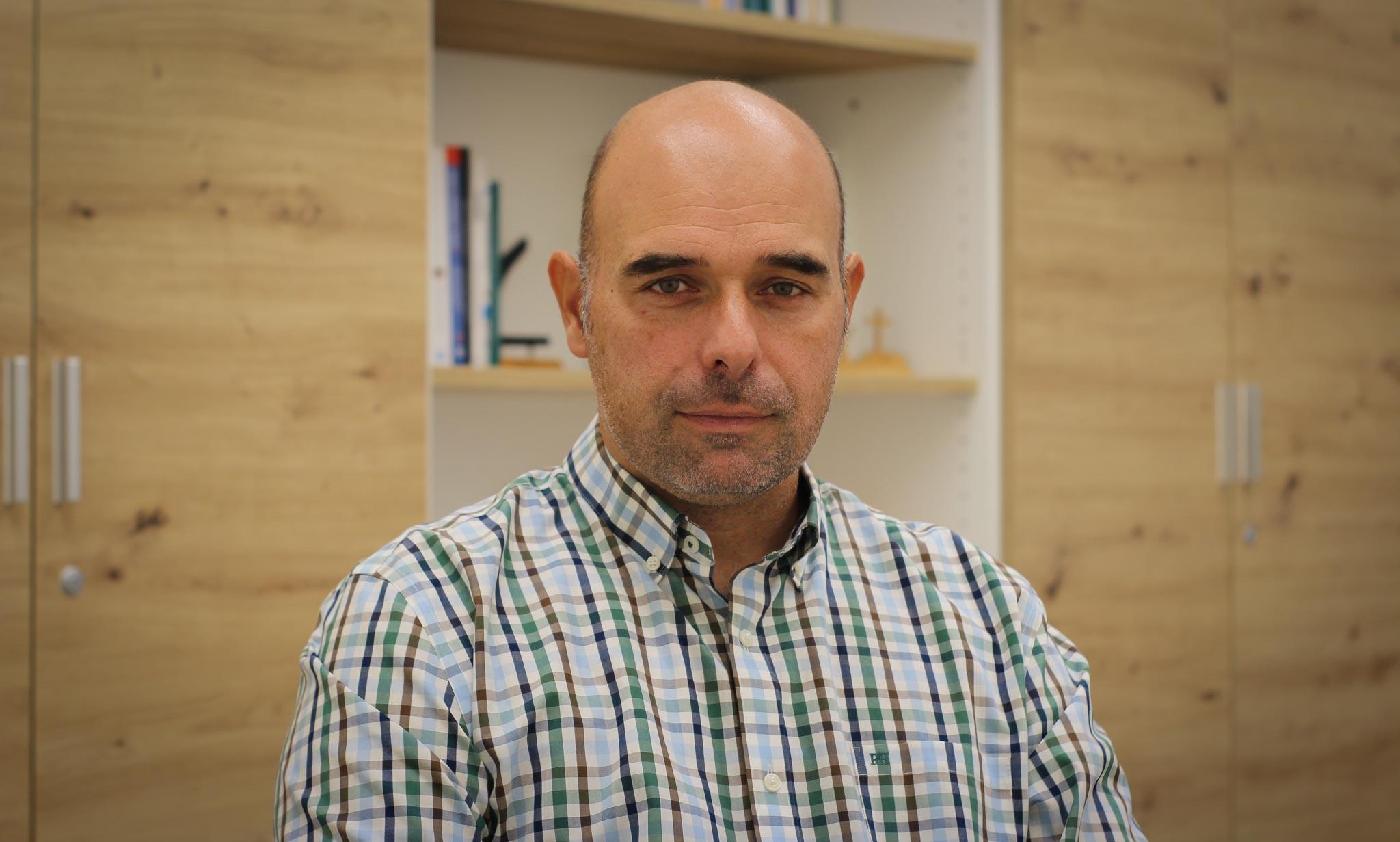 A bald man in a plaid shirt standing in front of a wooden bookshelf.