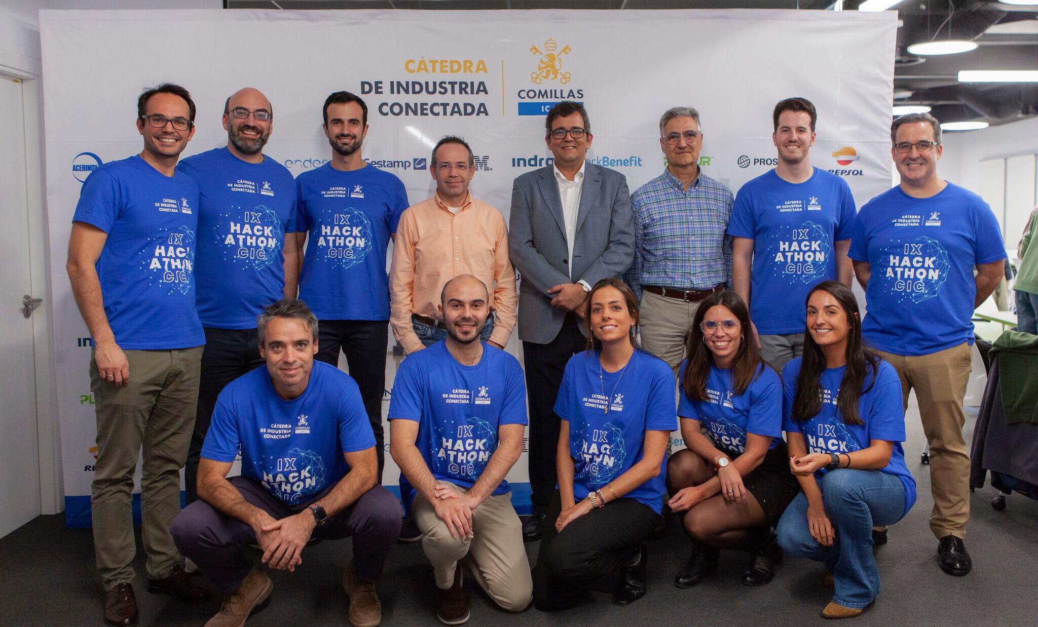 A group of people wearing blue Hackathon t-shirts, smiling for a photo in a room with a banner reading 'Cátedra de Industria Conectada Comillas'