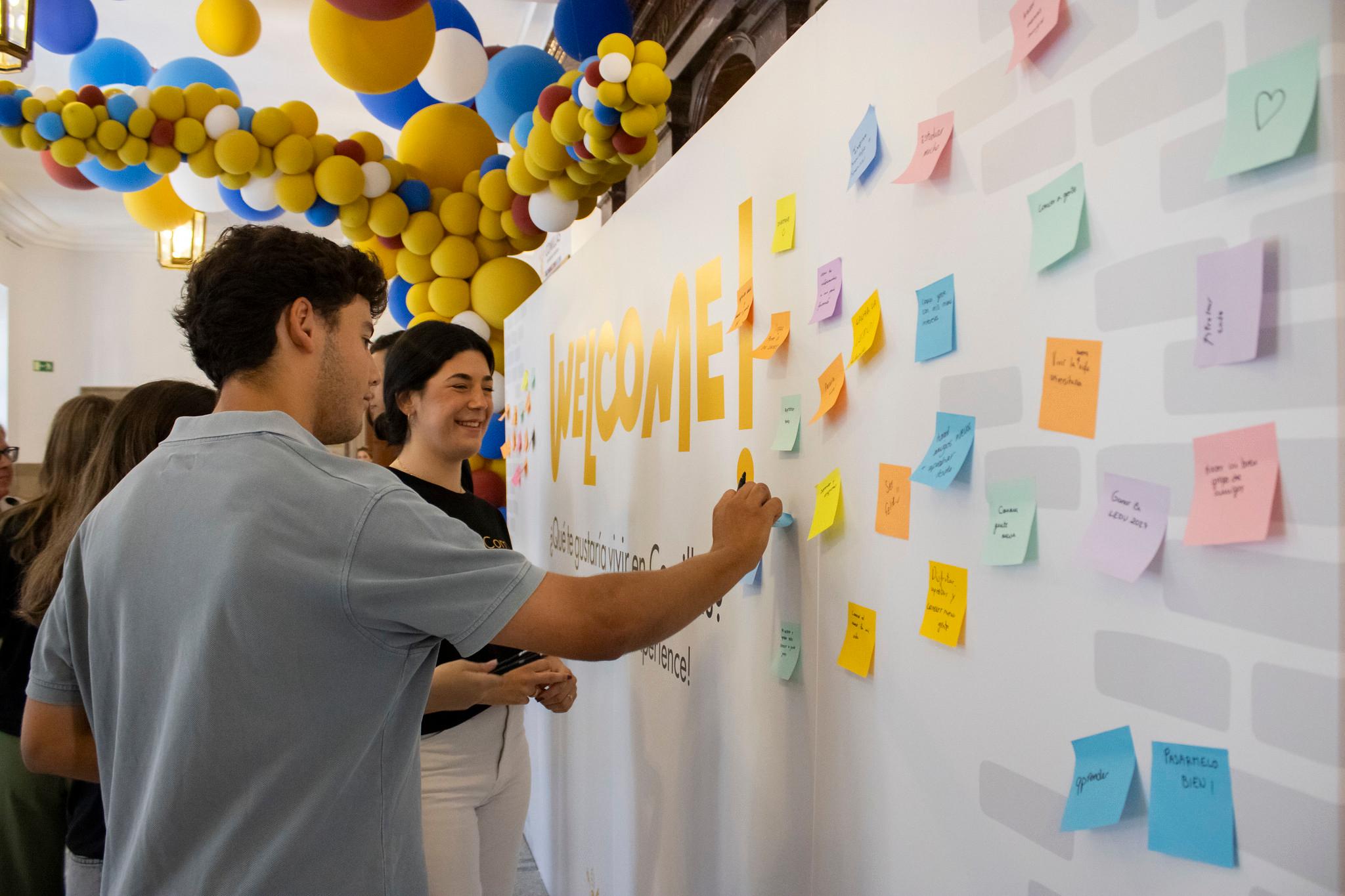 People placing sticky notes on a wall at a festive event with balloons and a 'Welcome' sign.