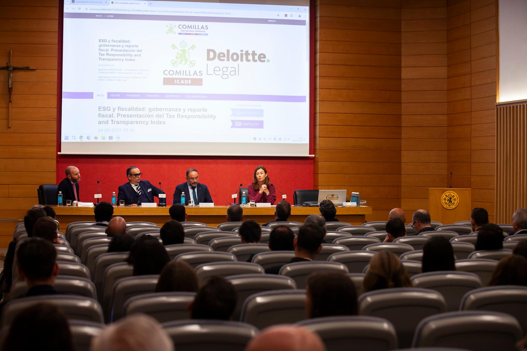 Four people participate in a panel discussion at a conference in an auditorium with audience members listening attentively.