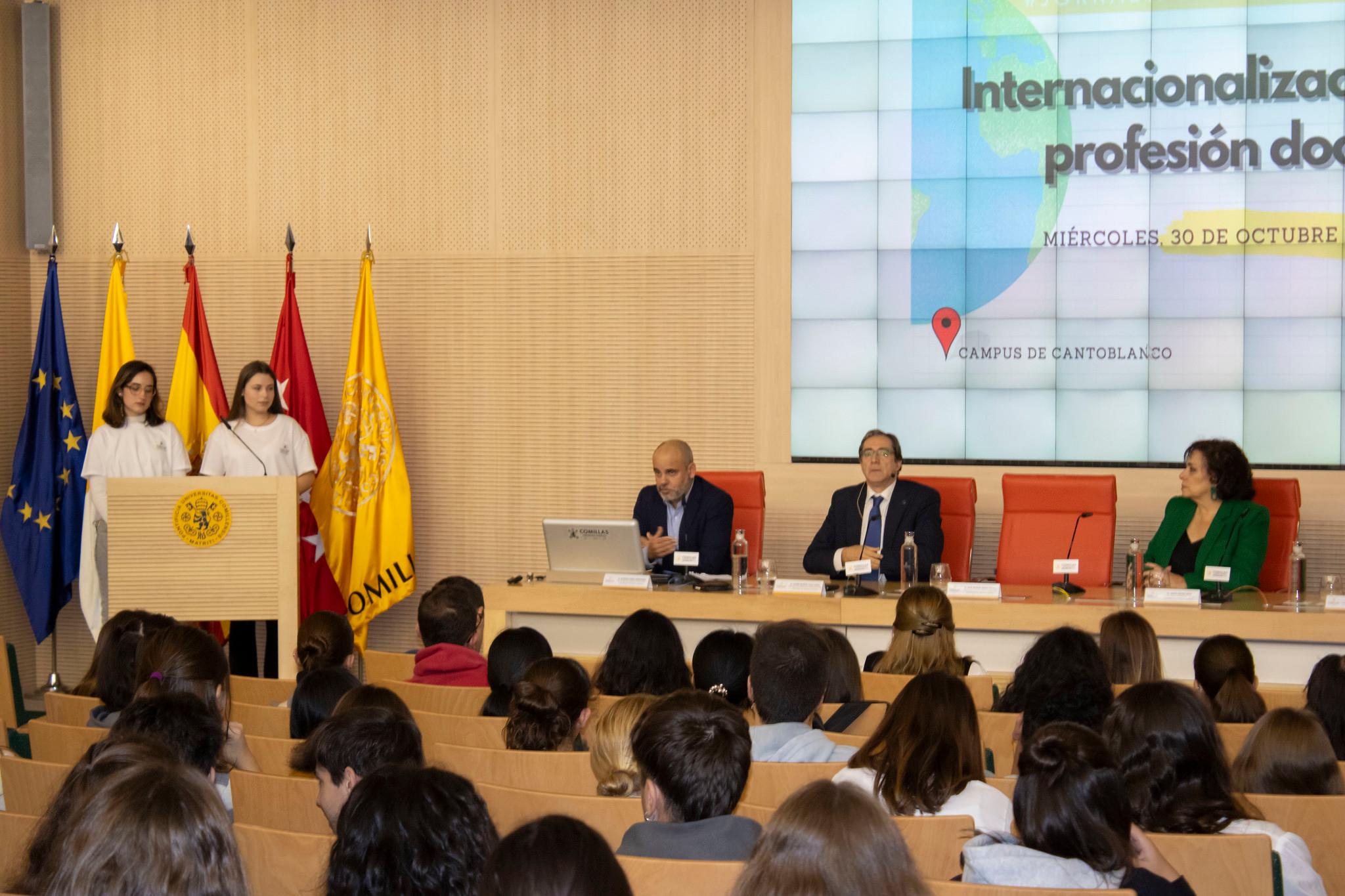 A group of people at a conference setting, with a speaker addressing an audience in a room with flags of the European Union and Spain.