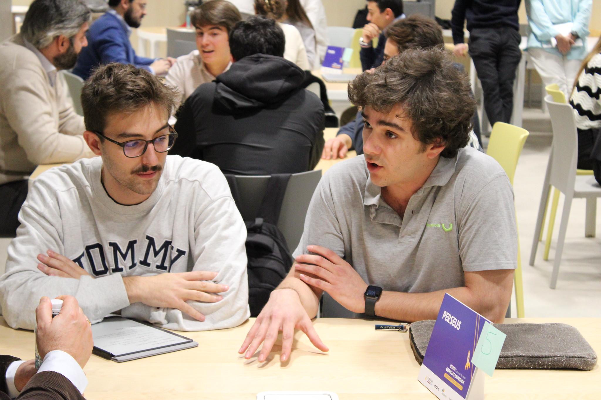 Dos hombres jóvenes conversando en una mesa con libros y documentos durante un evento o reunión.