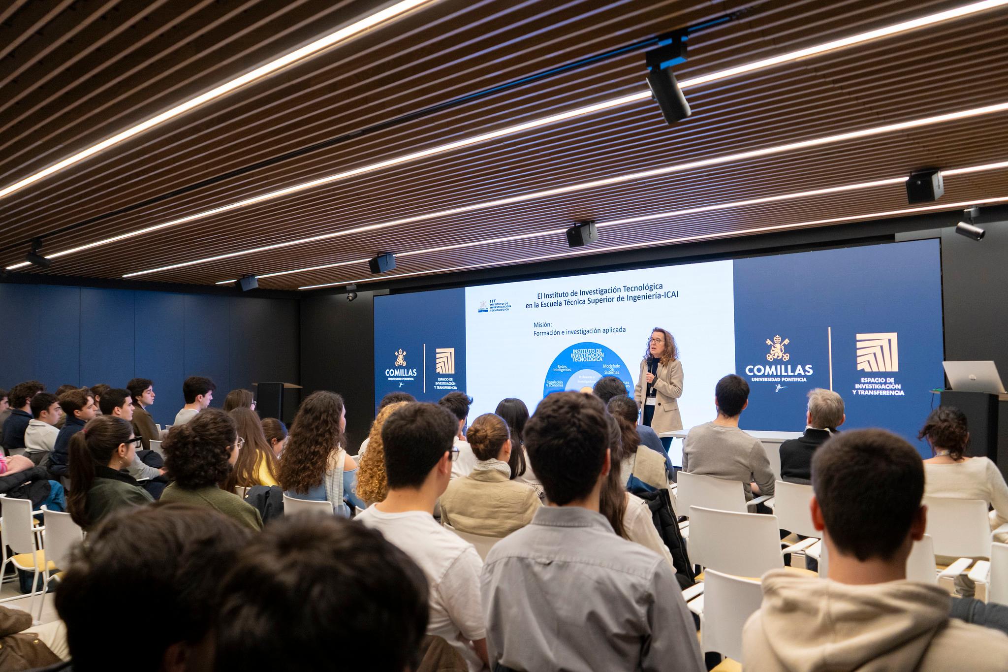 Una presentadora habla frente a un público joven en un evento sobre tecnología en un auditorio moderno.