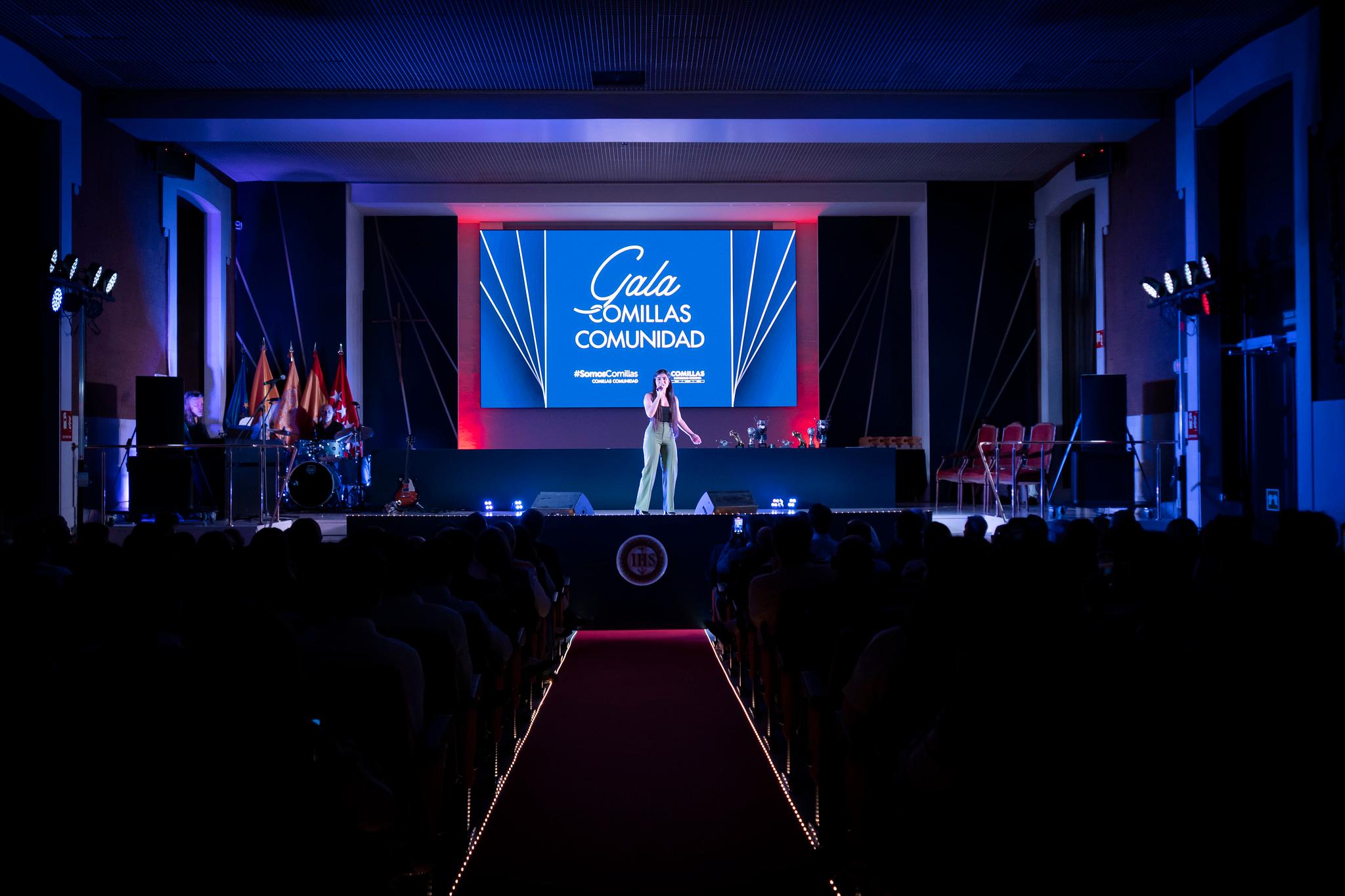 Una gala en un escenario iluminado, donde una persona se presenta frente a un público con un telón de fondo que dice 'Gala Comunidades'.