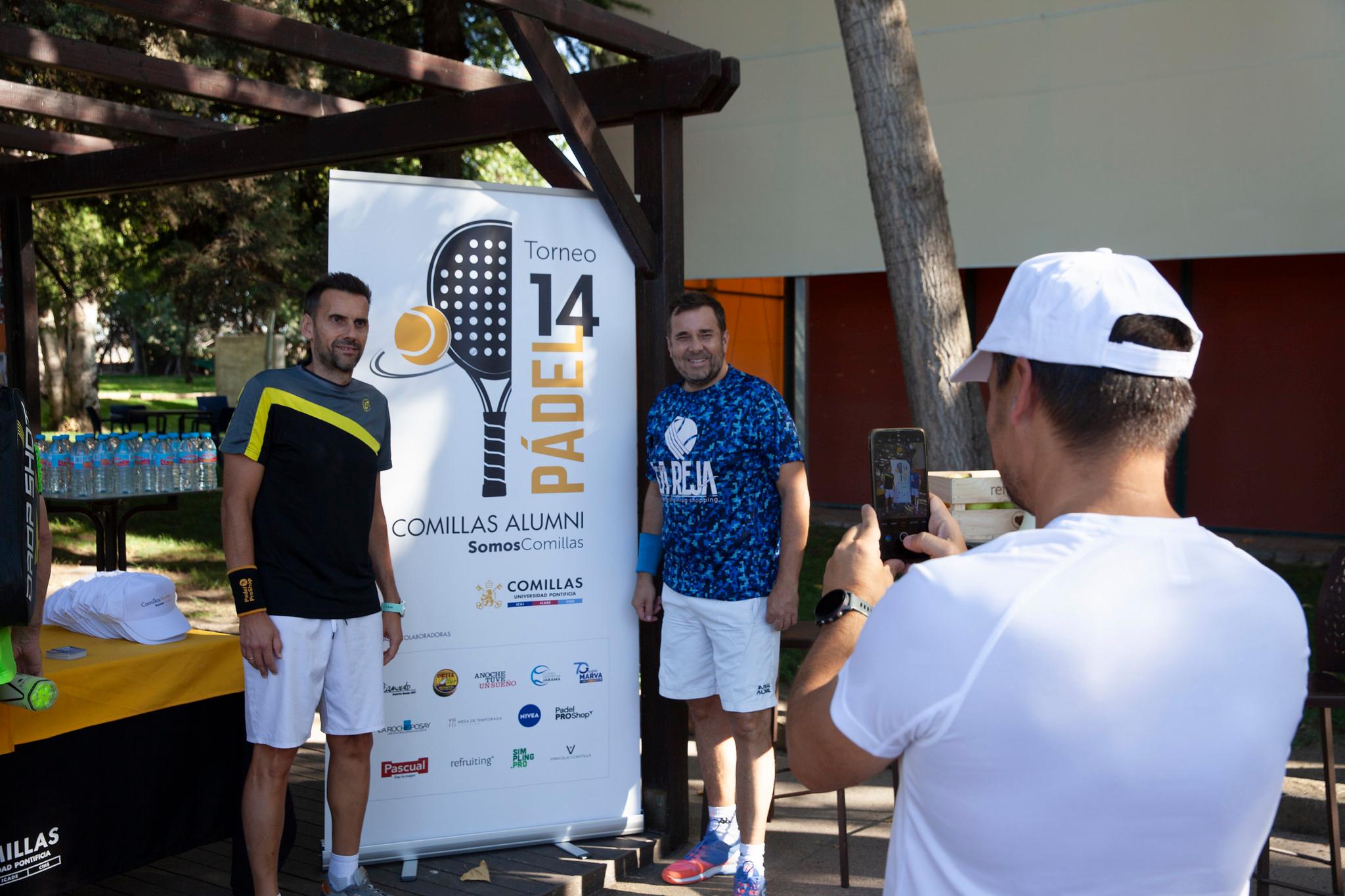 Dos jugadores de pádel posan frente a un cartel del Torneo 14 de Pádel Comillas Alumni mientras un tercero les toma una foto.