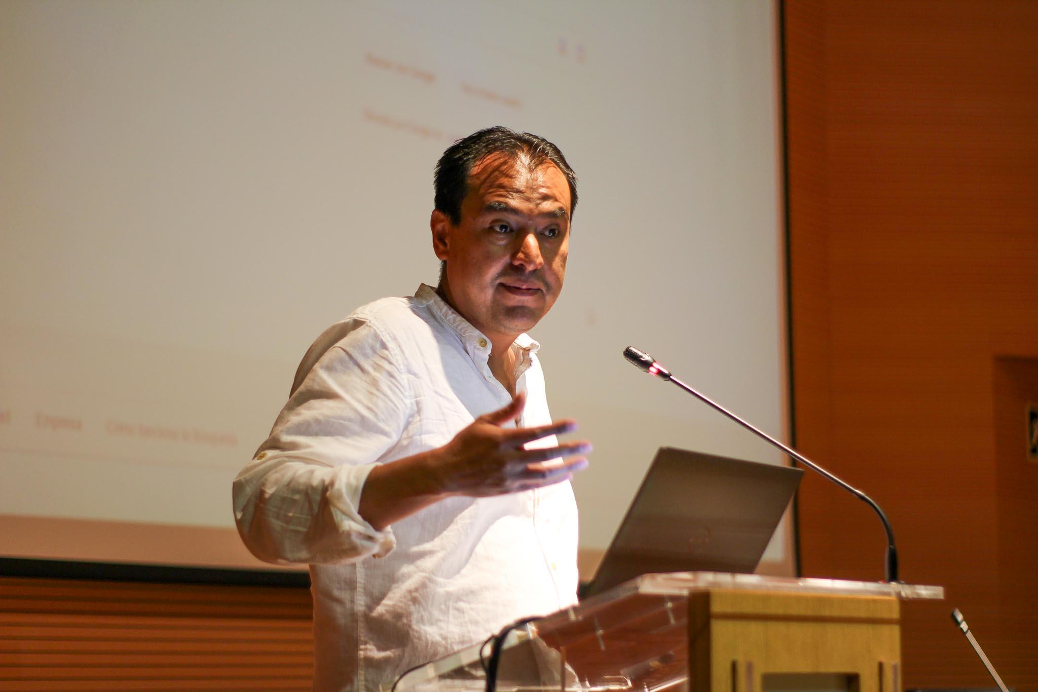 A man in a white shirt is giving a presentation at a podium with a projection screen in the background.