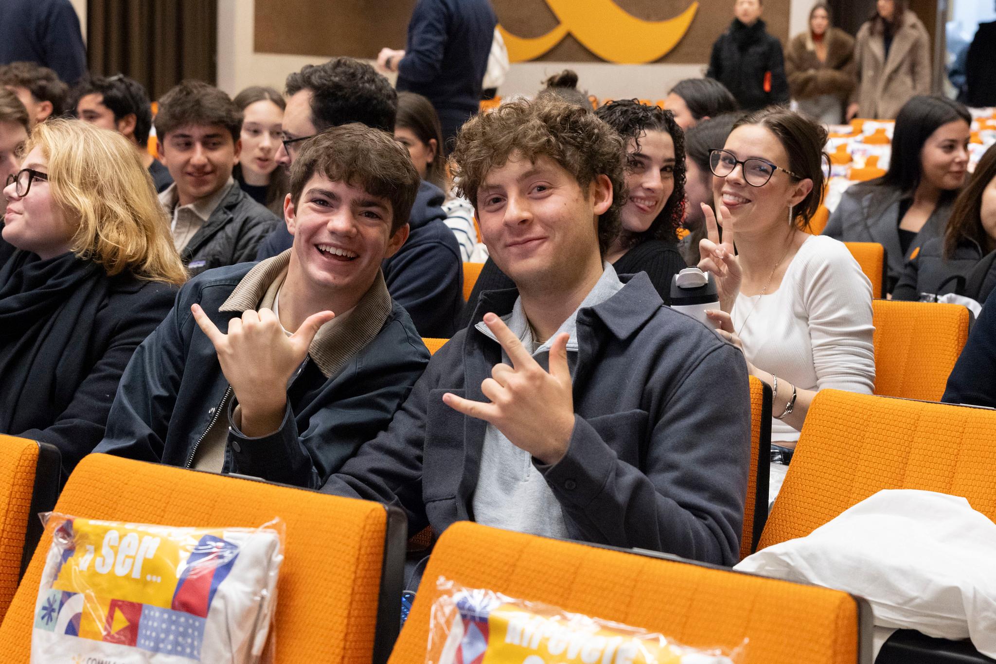 Un grupo de jóvenes sonrientes en un evento, sentados en una sala con asientos naranjas.