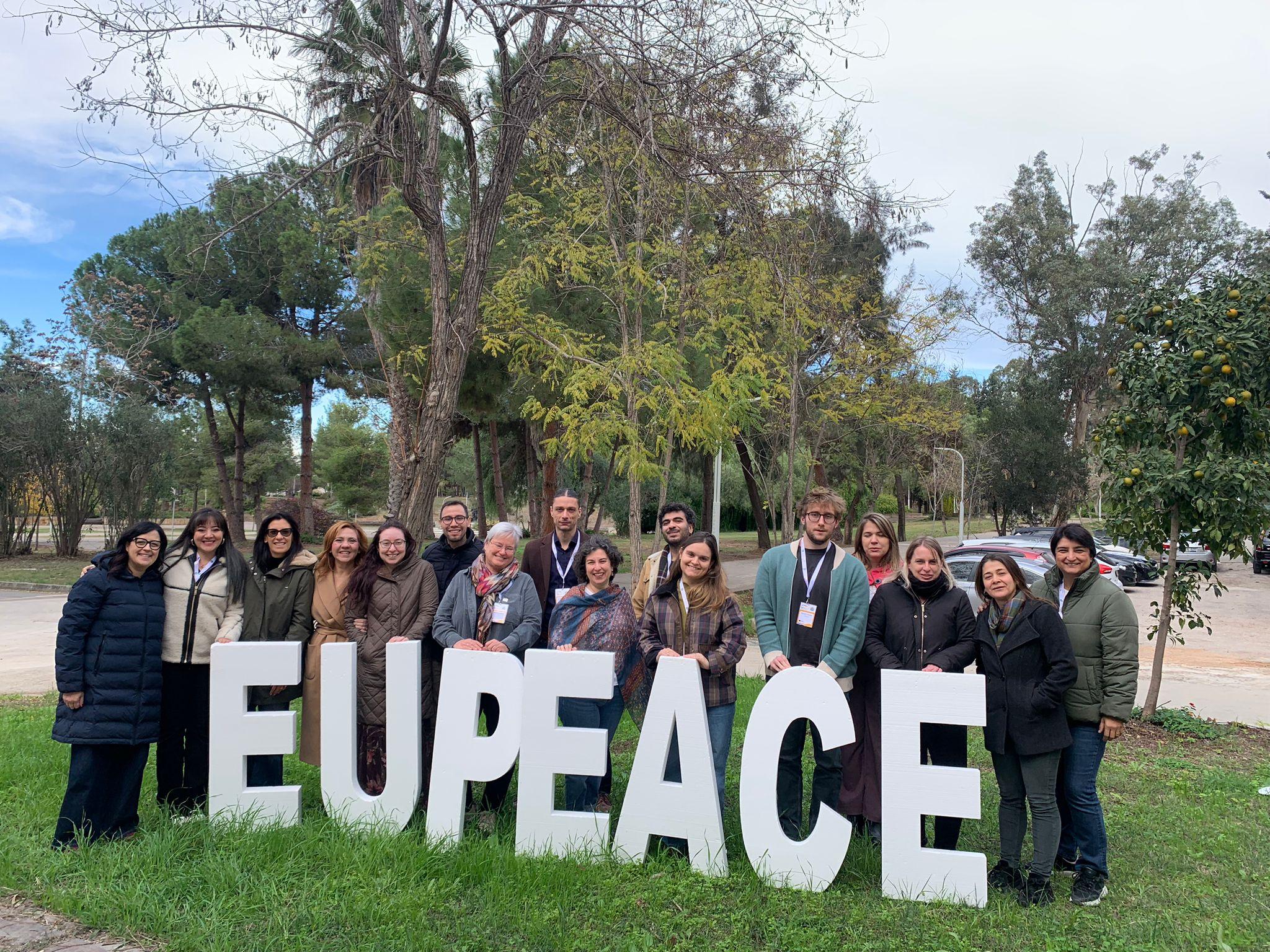 Un grupo de personas posa frente a un cartel grande que dice 'EUPEACE' en un entorno natural.