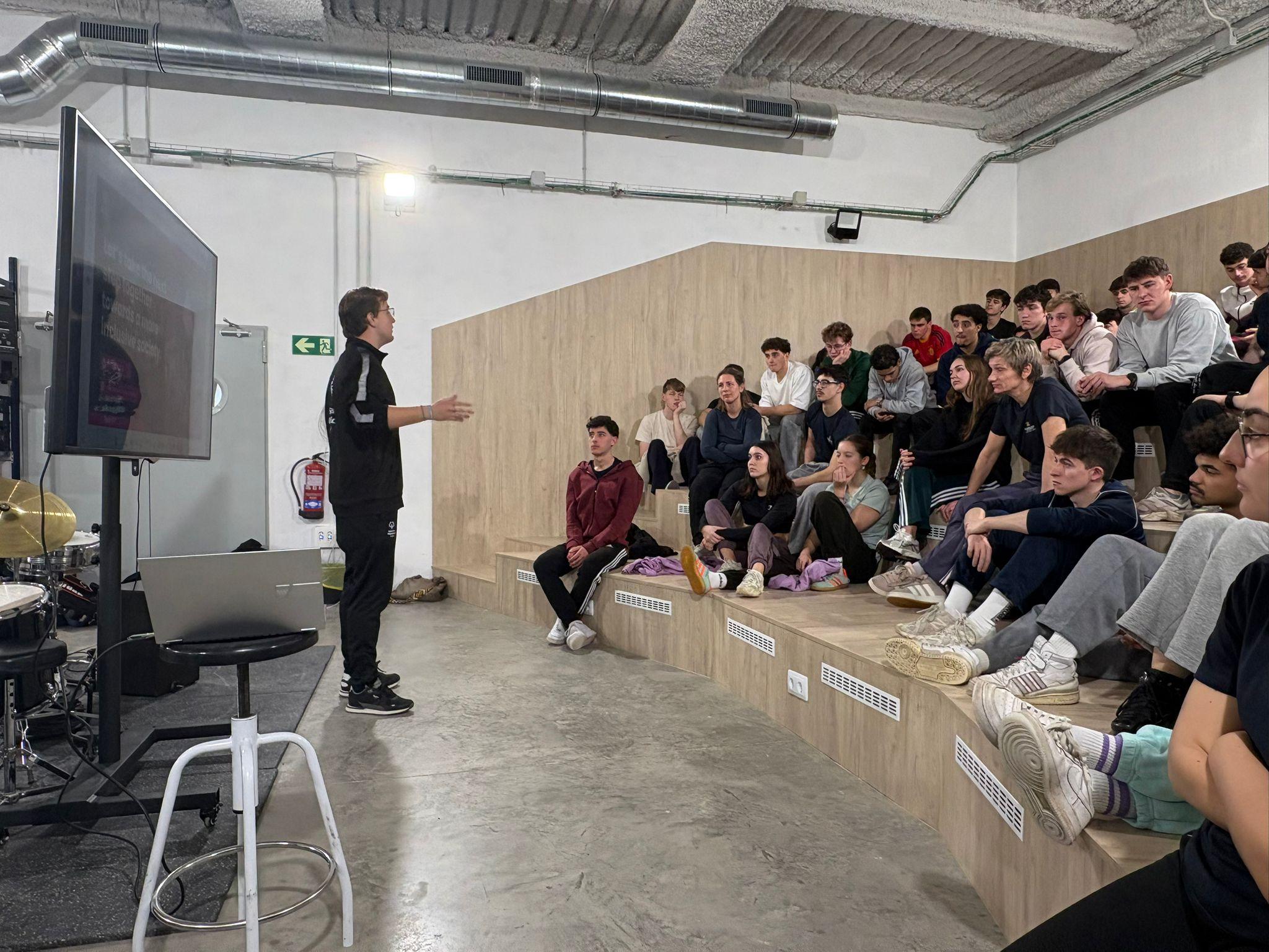 Un orador se dirige a un grupo de jóvenes sentados en gradas en un espacio moderno.