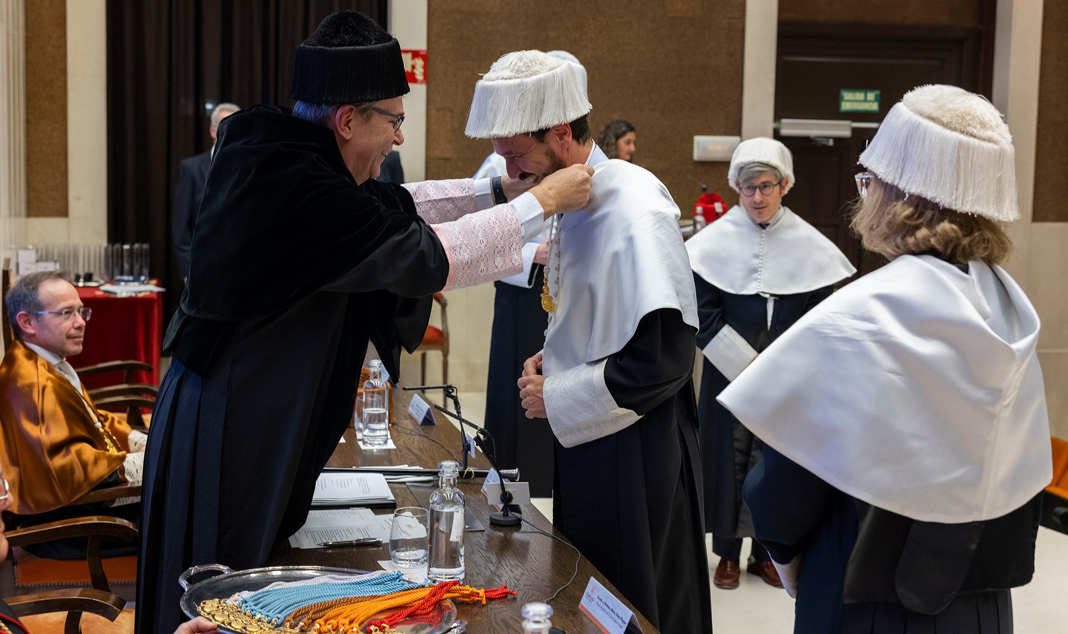 Una ceremonia académica en la que un hombre recibe un distintivo de manos de otro, mientras otros asistentes observan.