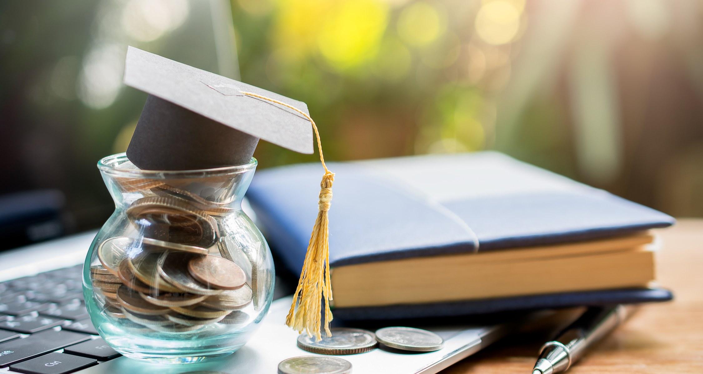 La imagen muestra una hucha con monedas y un gorro de graduación junto a un libro y un bolígrafo sobre una mesa.