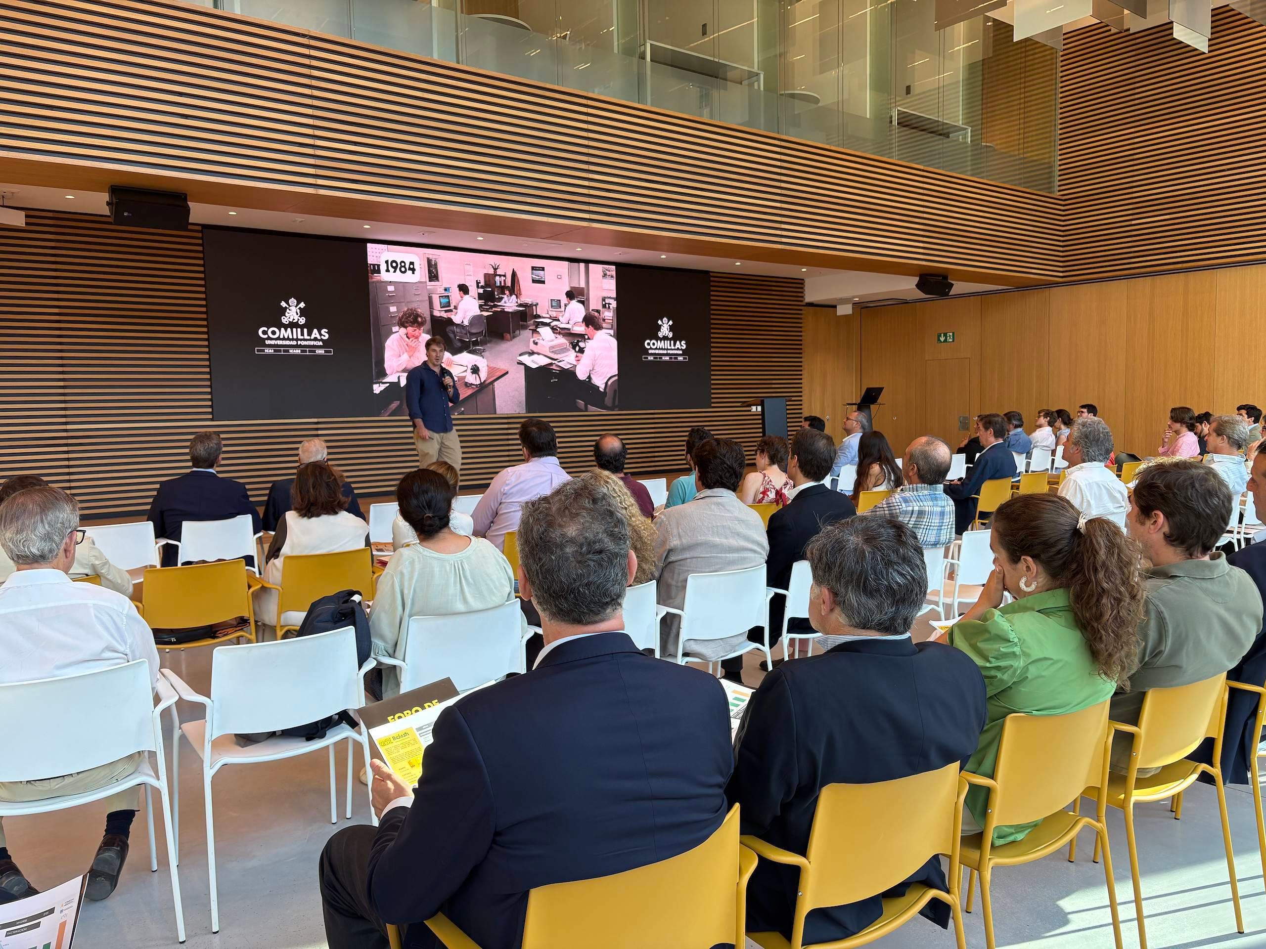 Una reunión celebrada en un auditorio moderno con un orador frente a una pantalla grande y una audiencia atenta.