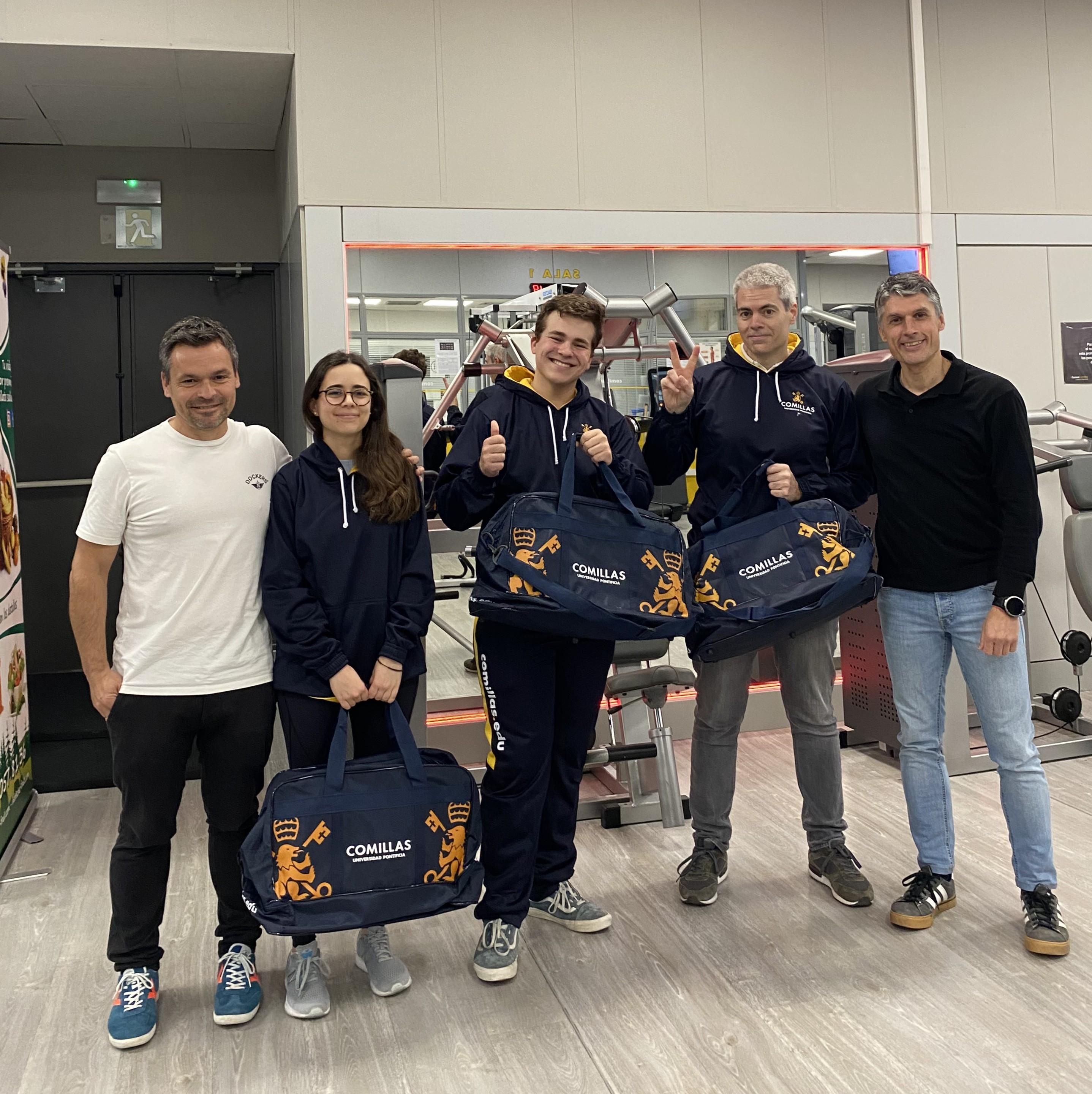 Un grupo de personas en un gimnasio sosteniendo bolsas con el logo de una institución.