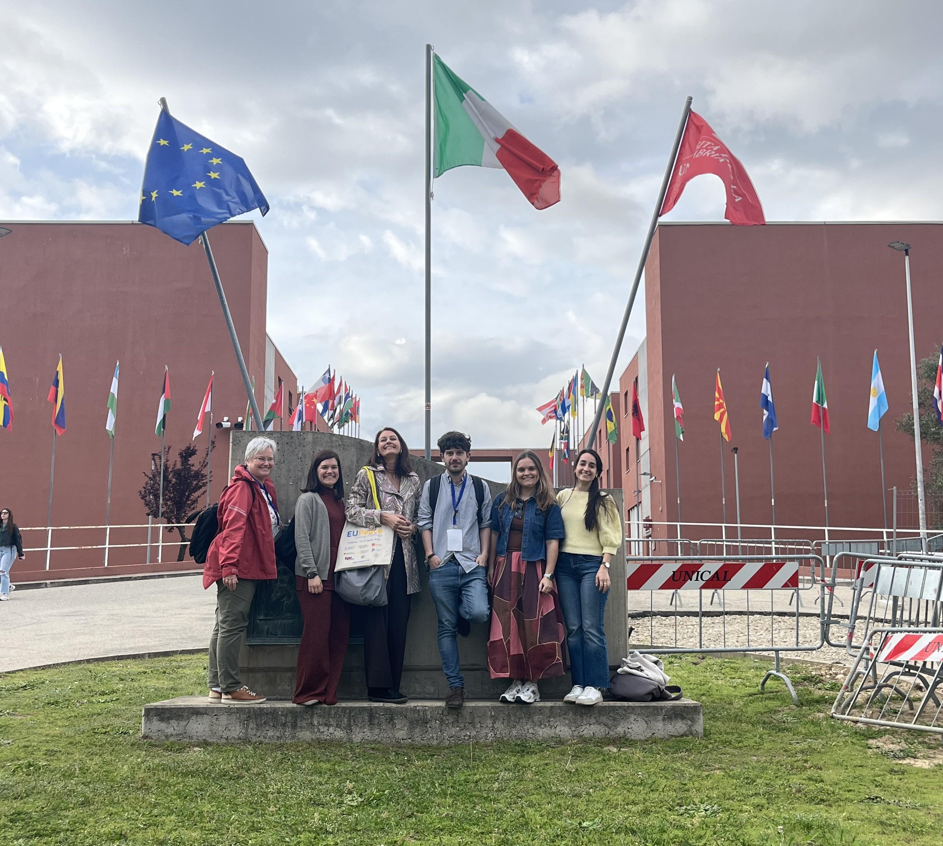 Un grupo de seis personas posan frente a varias banderas en un lugar al aire libre.