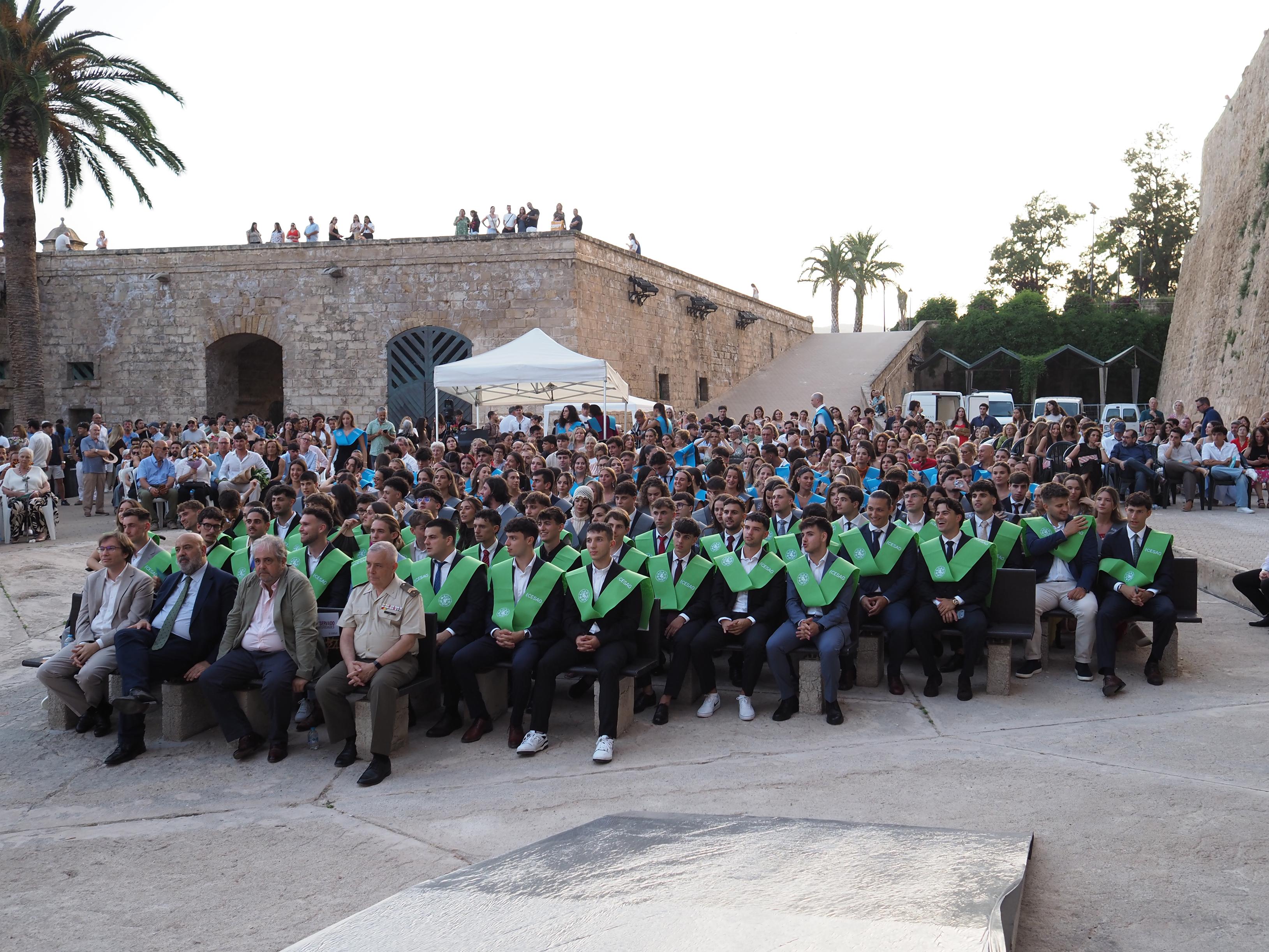 Un grupo numeroso de personas asistiendo a una ceremonia al aire libre, con una gran cantidad de graduandos en primera fila vistiendo togas con capes verdes.
