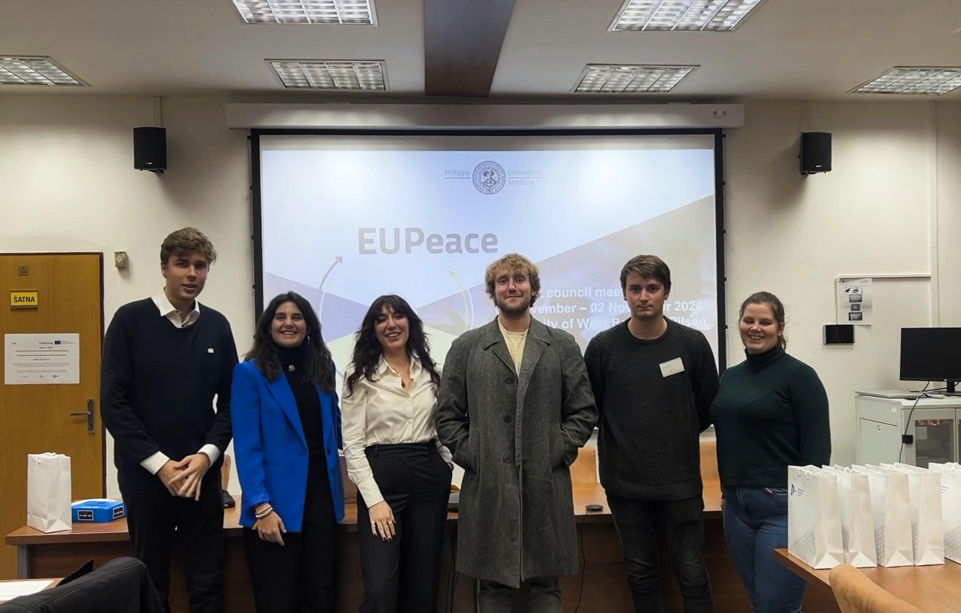 Grupo de seis personas jóvenes sonriendo en una sala de conferencias con una presentación de EUPeace en la pantalla.