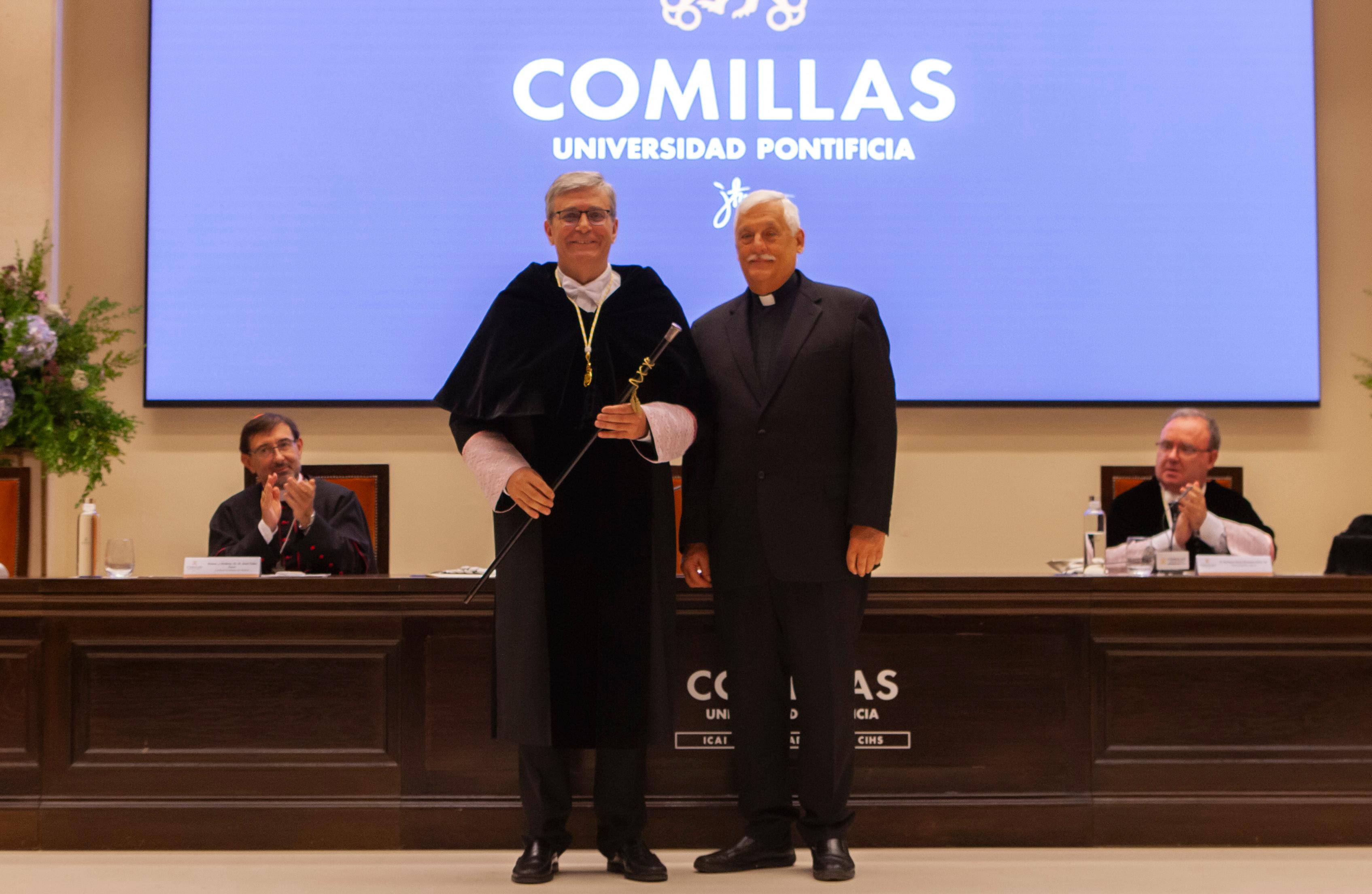 Two men, one in academic regalia, standing in front of a podium under a screen displaying 'COMILLAS Universidad Pontificia', shaking hands.