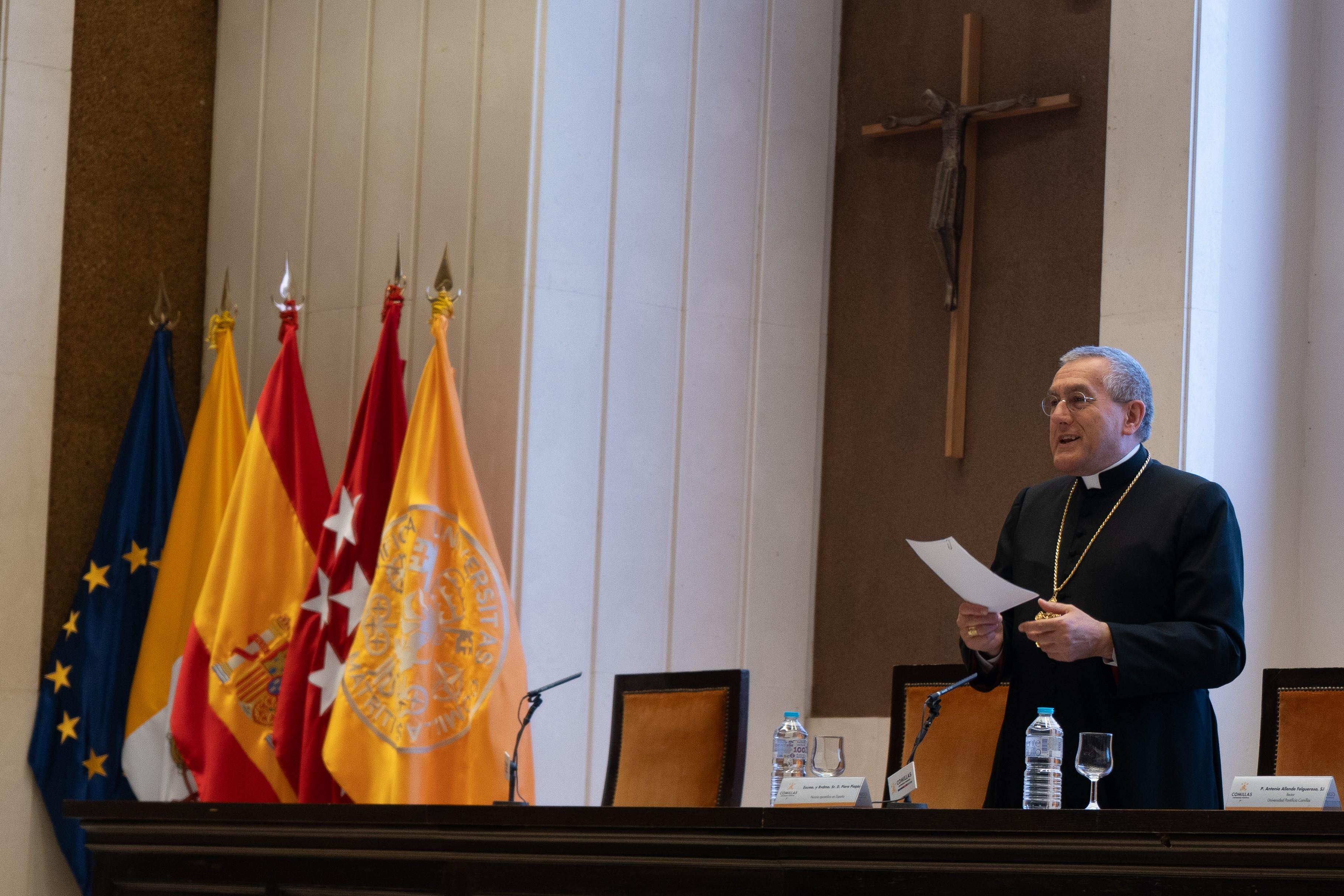 Un hombre vestido de clérigo se encuentra de pie frente a un atril, sosteniendo un documento, con banderas de España y una cruz en el fondo.