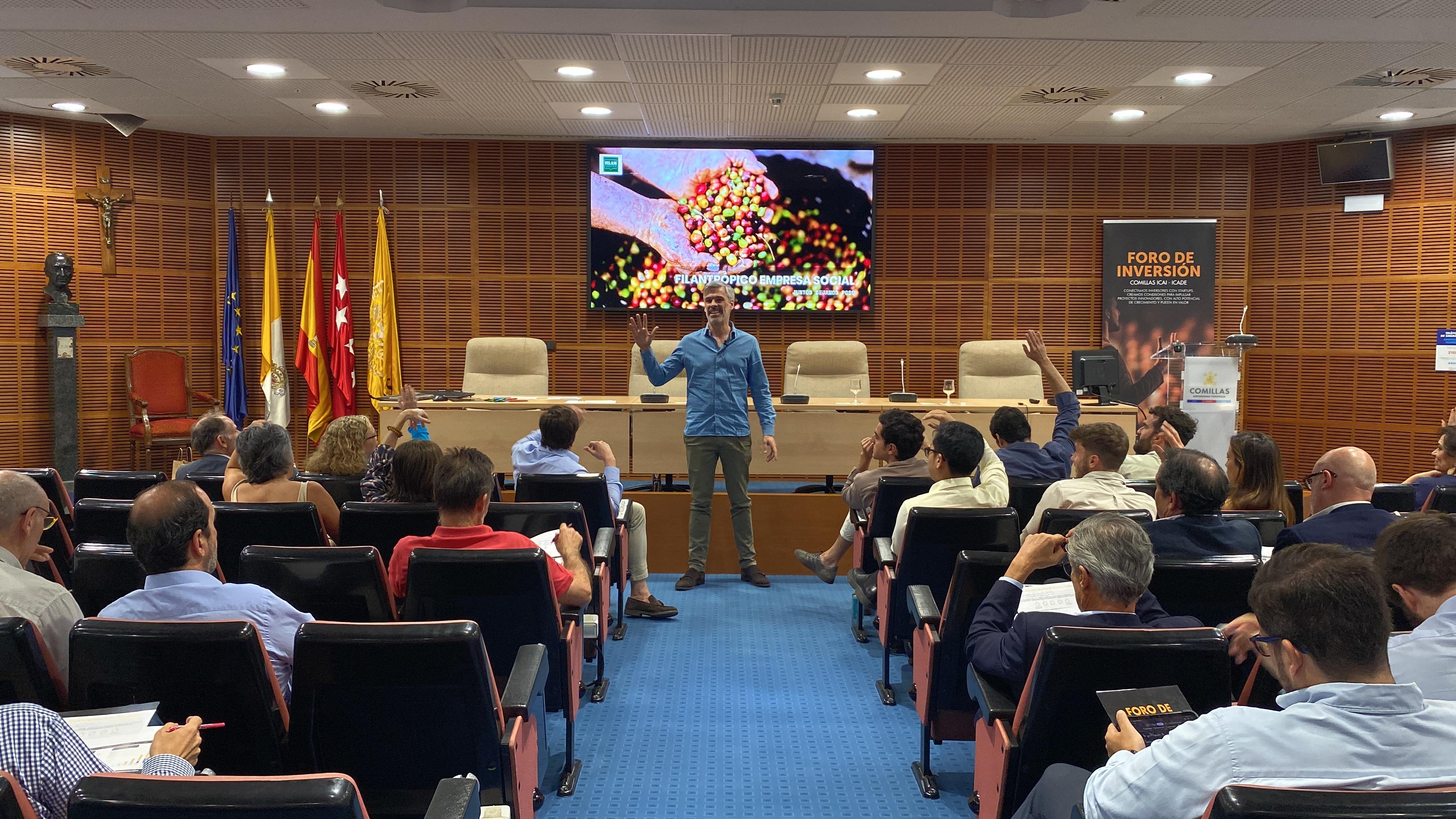 A speaker presenting at a conference in a room with an audience listening attentively, flags are displayed in the background.