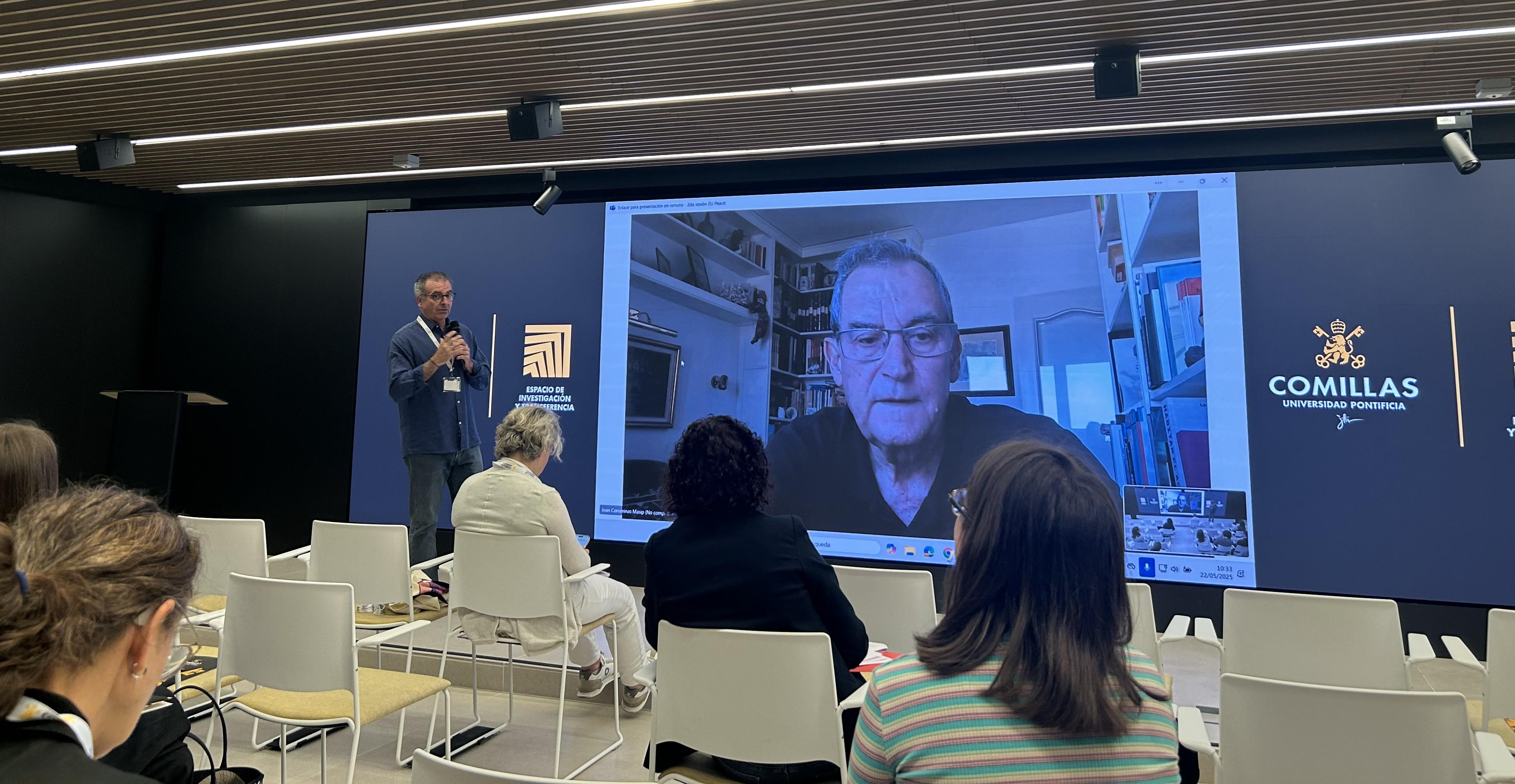 Un ponente está hablando frente a una pantalla en un evento en la Universidad Comillas, mientras un conferencista aparece por videollamada.