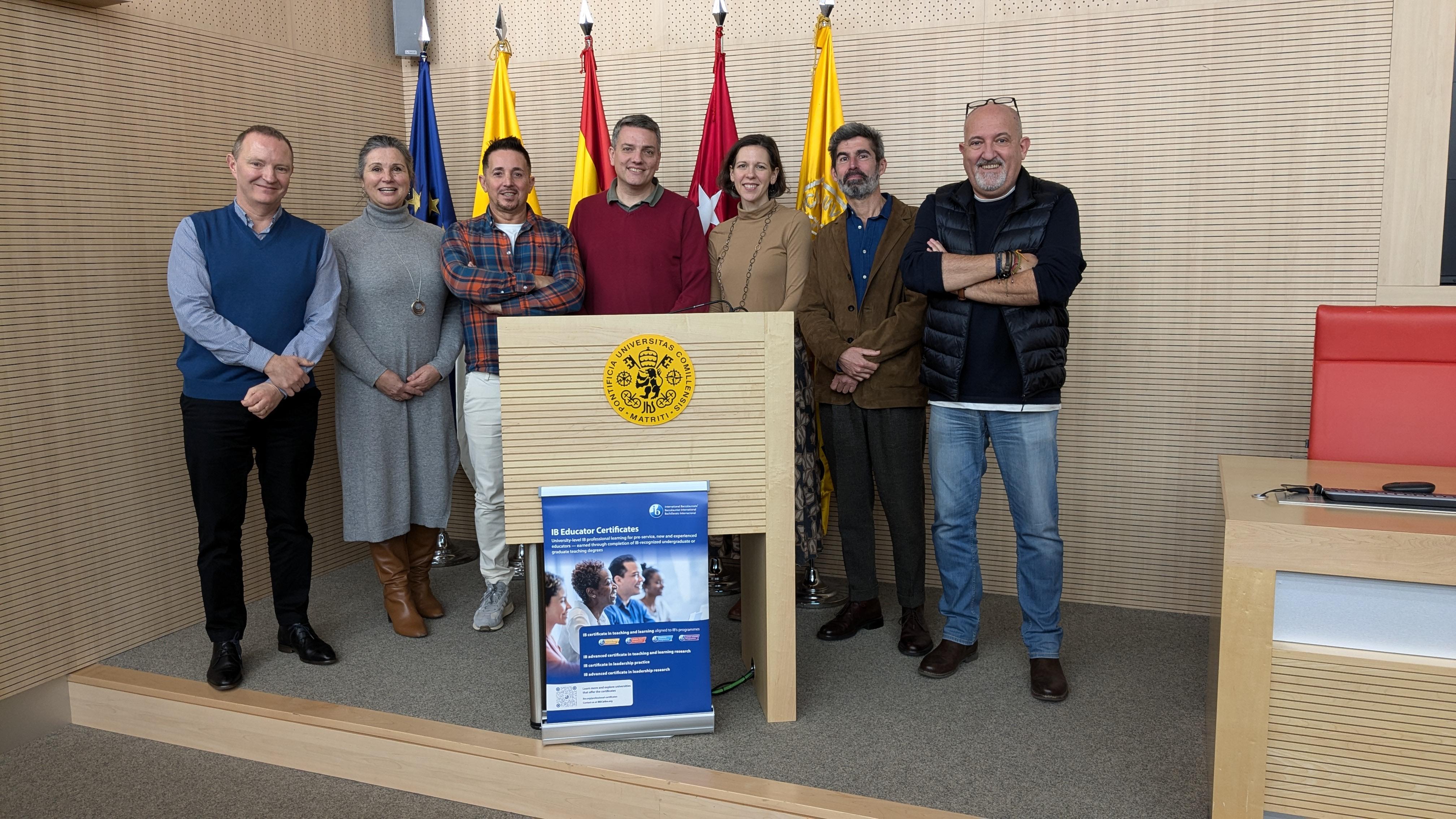 Grupo de siete personas posando en una sala con banderas y un cartel informativo.