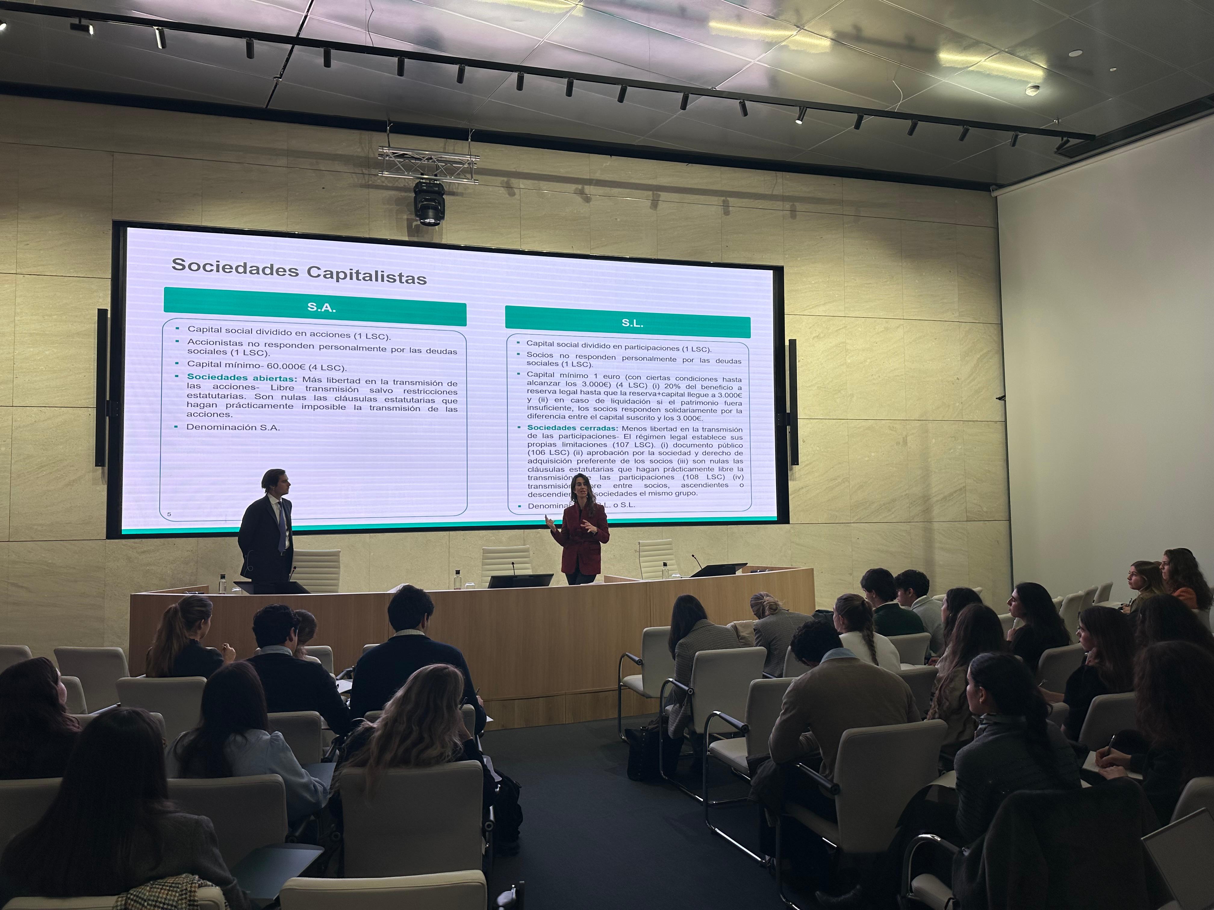 Una presentación sobre sociedades capitalistas se lleva a cabo frente a una audiencia en un salón moderno.