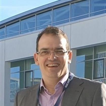 A smiling man in glasses stands outside a building, wearing a suit with a badge.