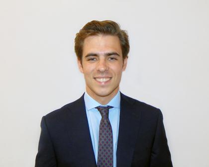 A young man in a blue suit and tie smiling in front of a plain background.