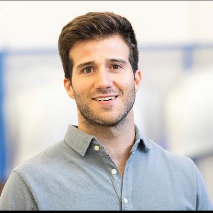 Smiling man with dark hair wearing a blue button-down shirt in an office setting.