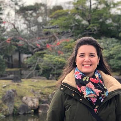 A woman smiling in a park with trees and a stone structure in the background.