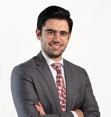A professional portrait of a smiling man wearing glasses, a gray suit, and a red tie with a playful pattern.