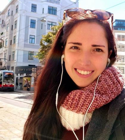 A smiling woman with a pink scarf and sunglasses on her head, wearing earphones in an urban setting.