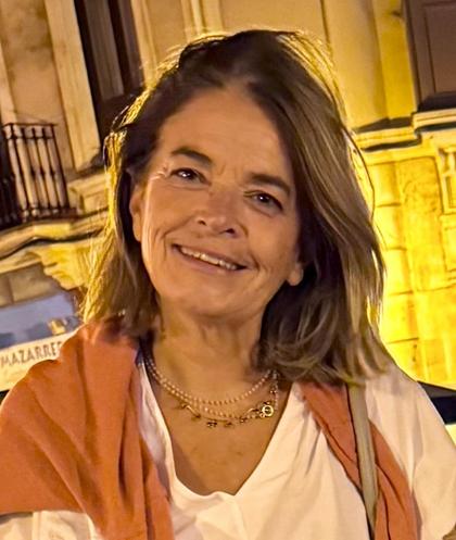 Una mujer sonriente, con cabello despeinado y vestida con una camiseta blanca y una chaqueta anaranjada, se encuentra en una calle iluminada de noche.
