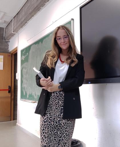 A woman standing in a classroom holding a book, smiling at the camera.