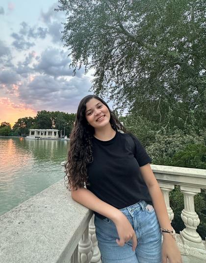 A young woman smiling by a lake with a sunset and trees in the background.