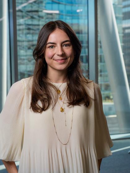 Una mujer sonriente con cabello largo, vestida con una blusa blanca, posa frente a grandes ventanales en un entorno urbano.
